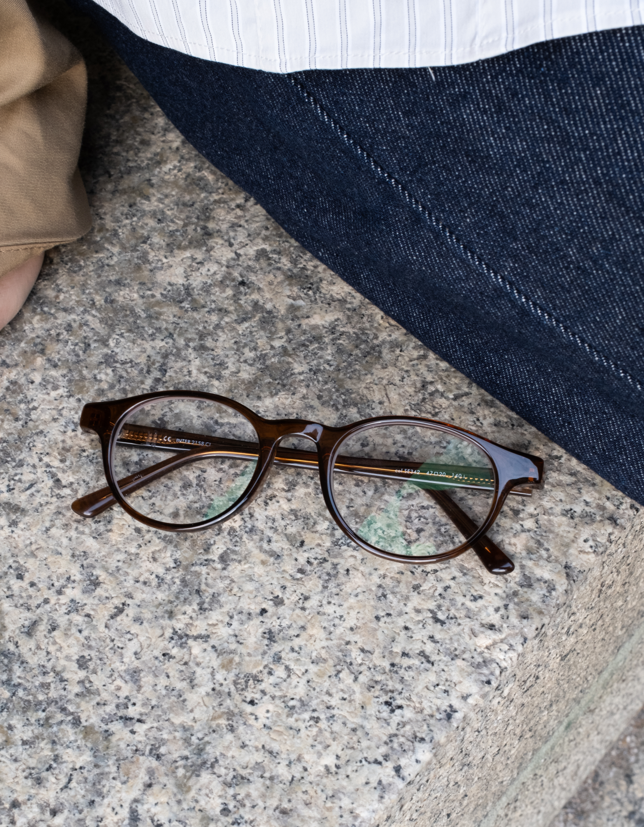 Occhiali rotondi marroni con lenti trasparenti poggiano su una superficie di pietra accanto a una persona che indossa jeans scuri e una camicia bianca.