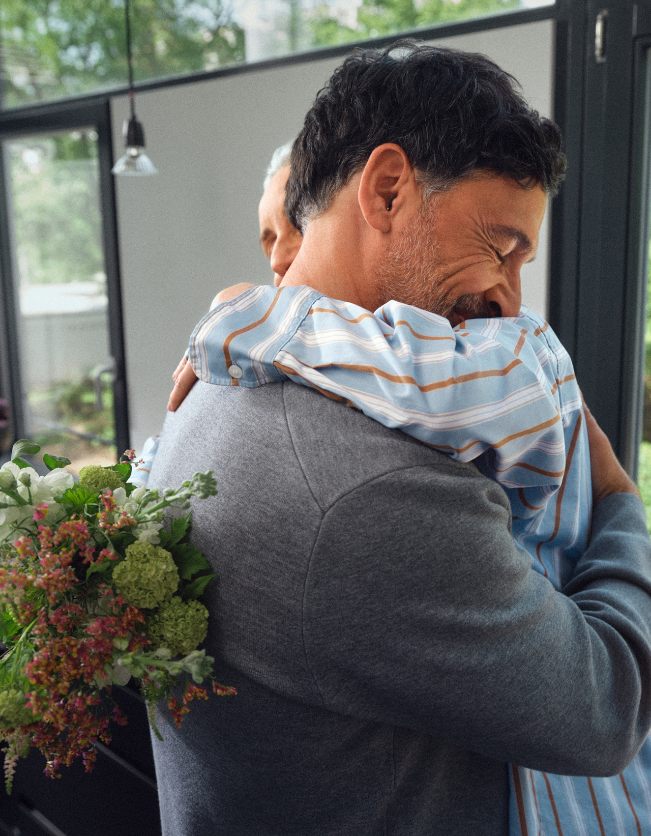 Ein Mann in einem grauen Pullover umarmt eine Person in einem gestreiften Hemd und lächelt herzlich.