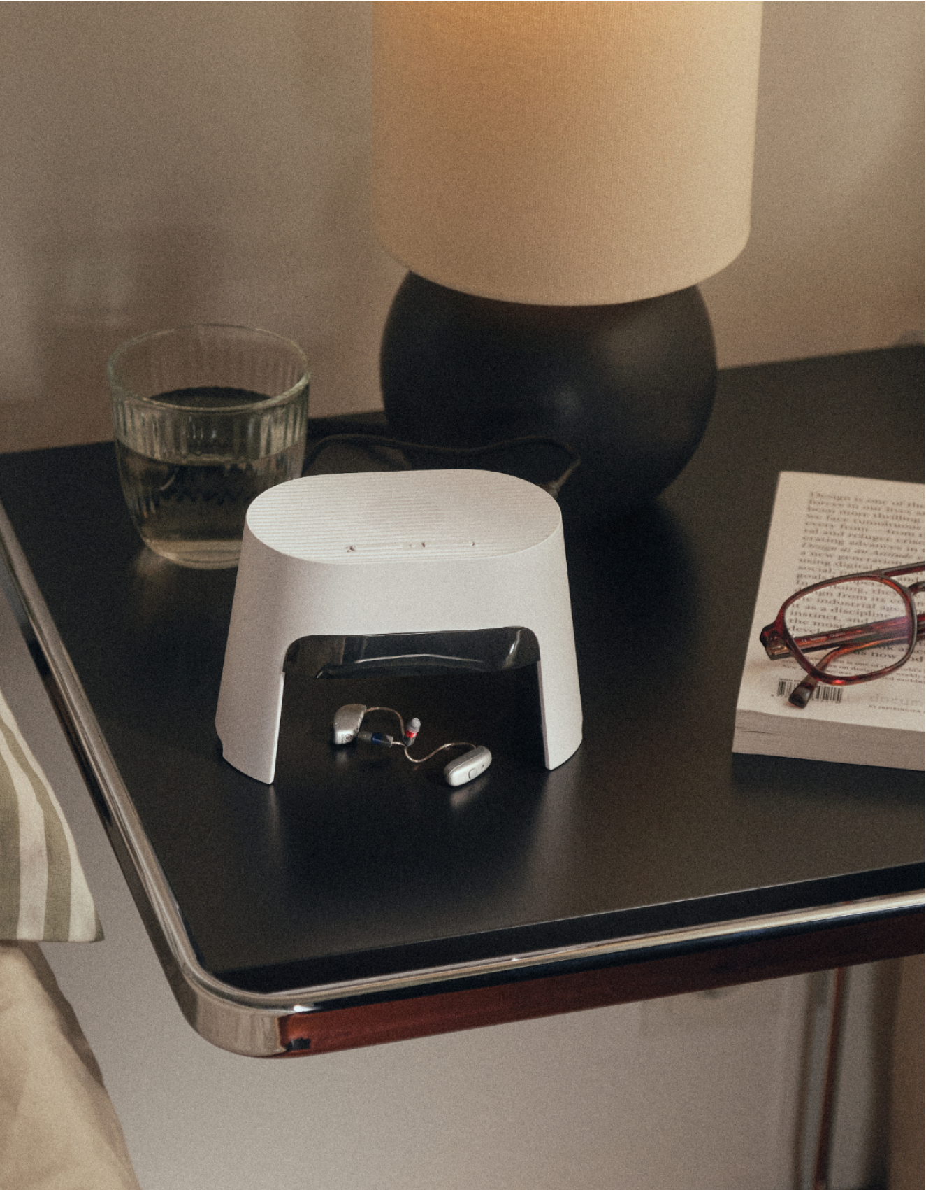Ein Nachttisch mit einer Lampe, einem Glas Wasser, einem weißen Gerät mit Hörgeräten drin, einem Buch und einer Brille.