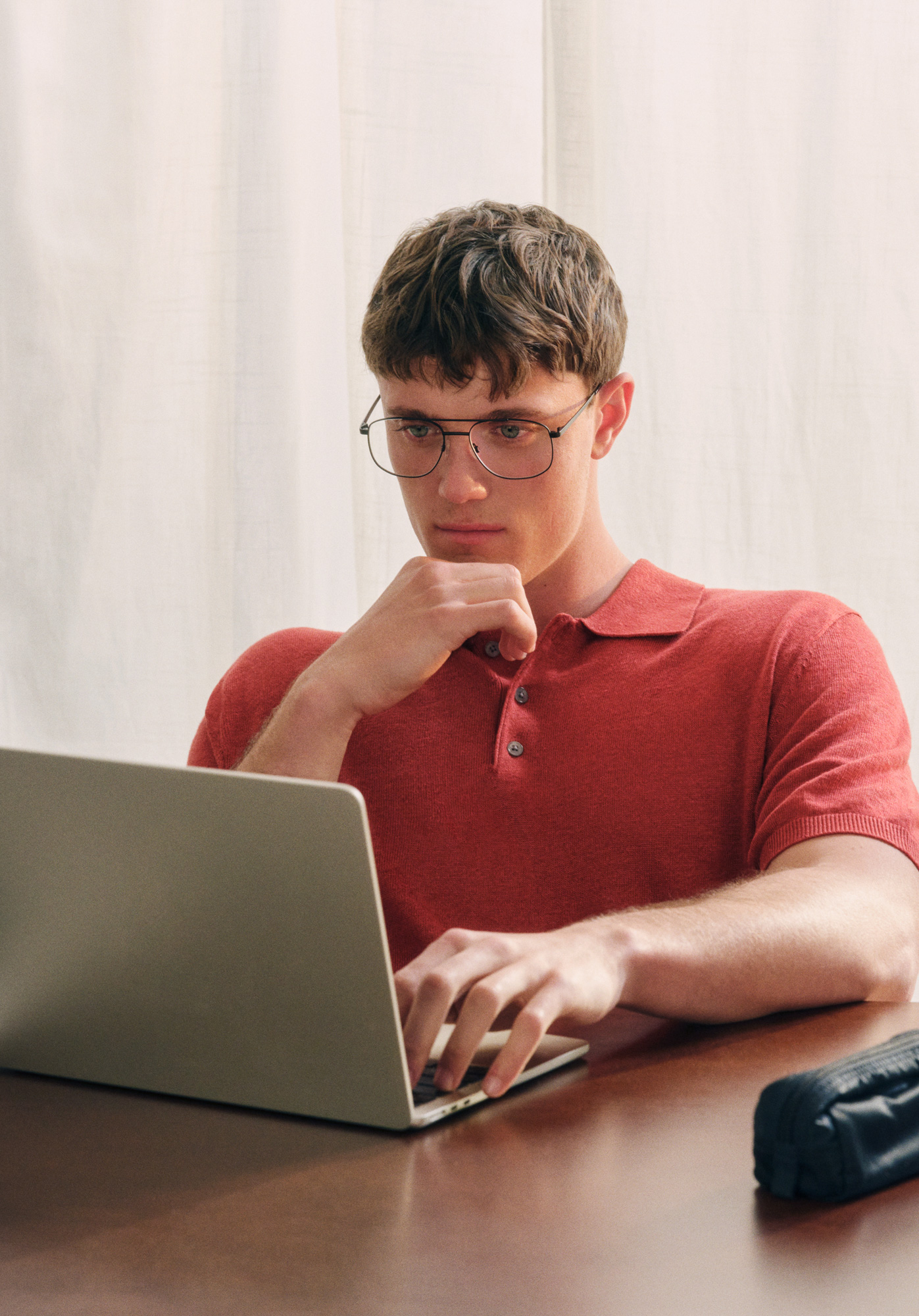 Eine Person mit Brille und rotem Polohemd arbeitet mit konzentriertem Gesichtsausdruck an einem Schreibtisch an einem Laptop.