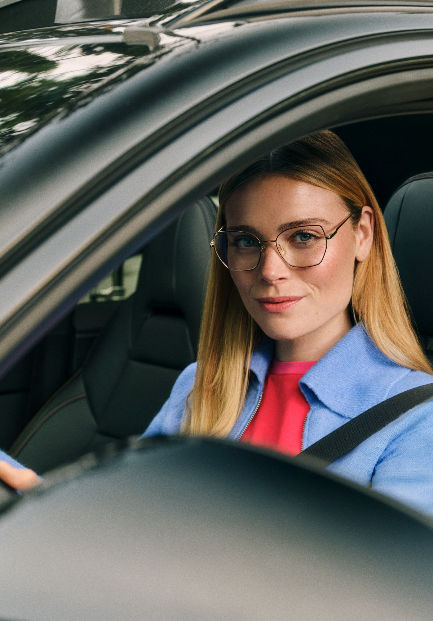 Una donna con gli occhiali e i capelli biondi, che indossa una giacca blu, è seduta al posto di guida di un'auto, guarda direttamente nella fotocamera ed è circondata da alberi.