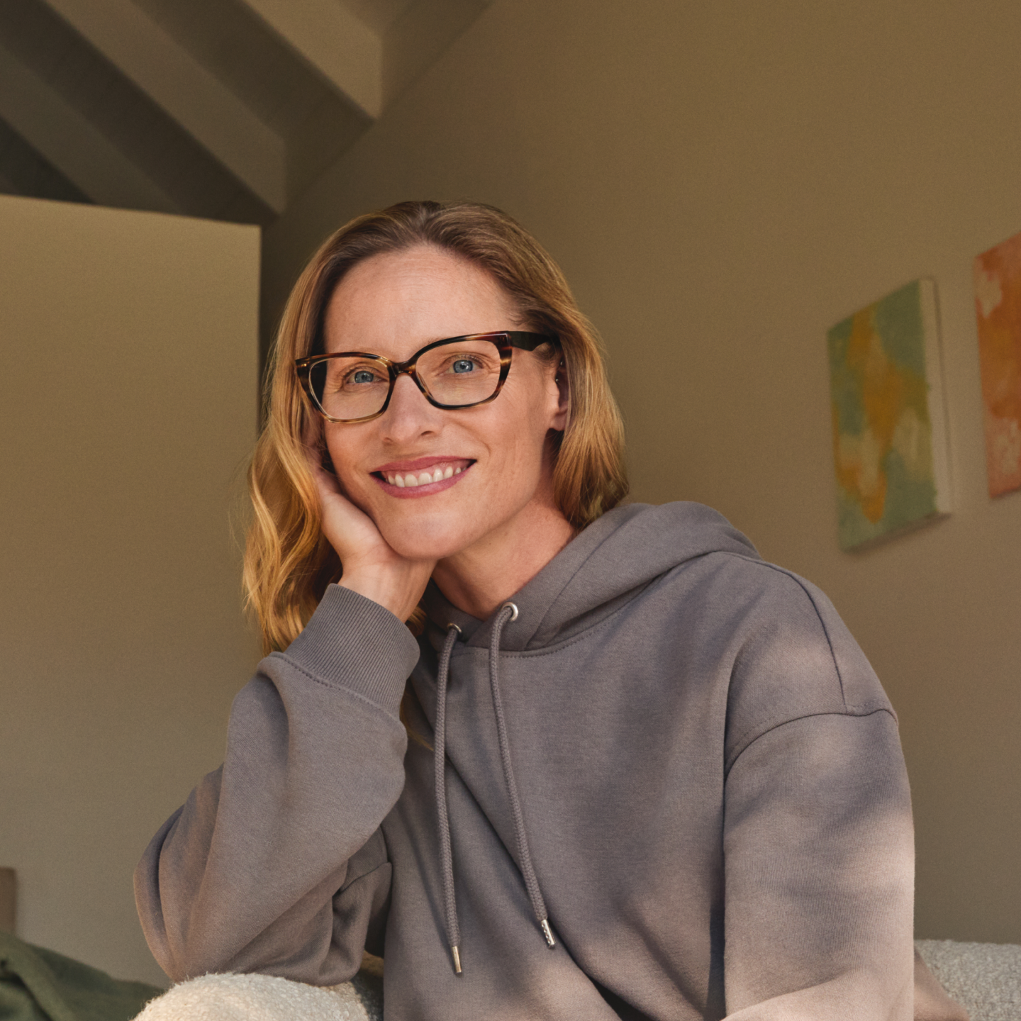 Lächelnde Frau mit Brille und grauem Kapuzenpulli, die ihren Kopf auf die Hand stützt und in einem Raum mit sanfter Beleuchtung und farbenfrohen Wandbildern im Hintergrund sitzt.