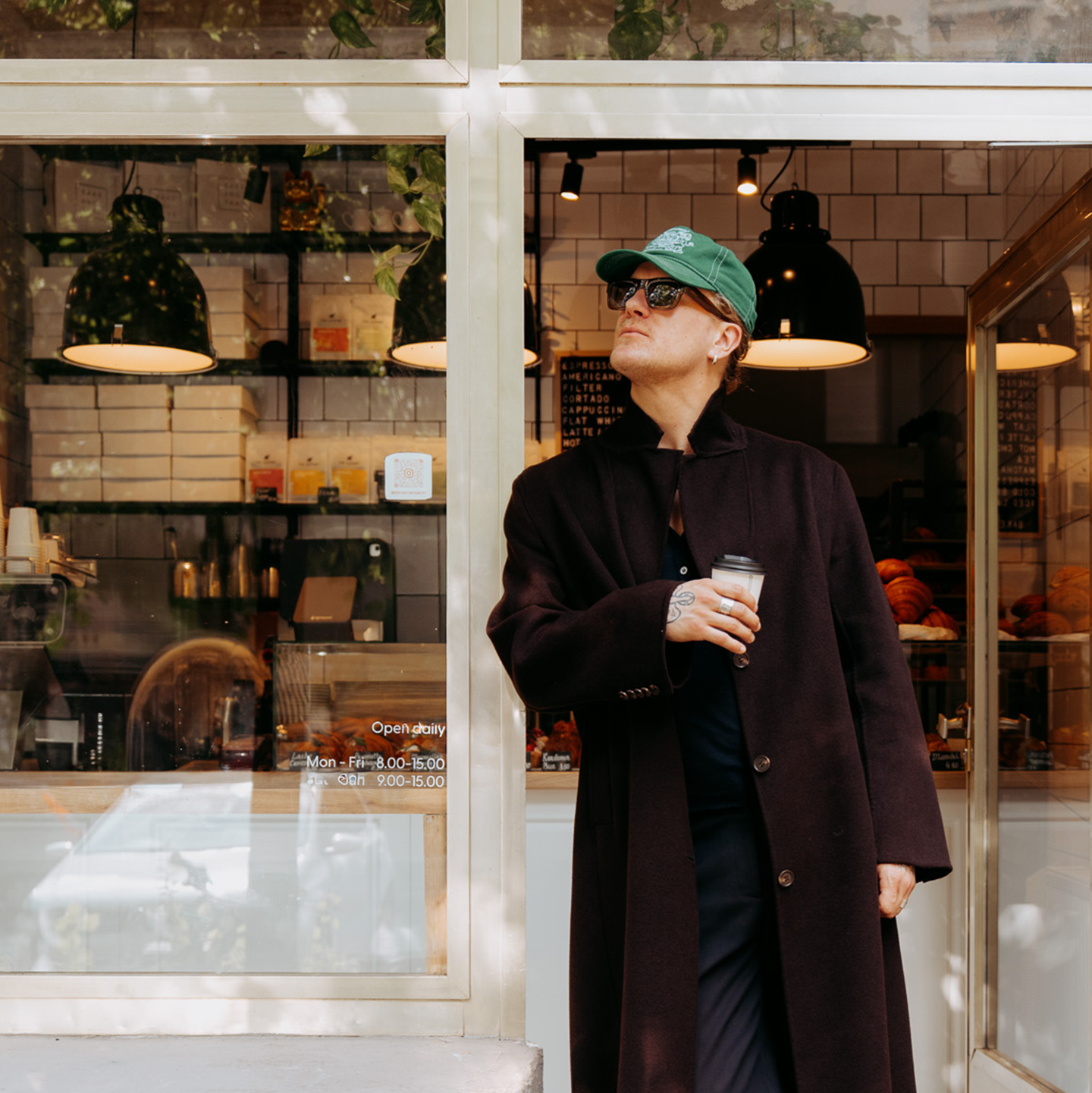 L’artiste Peter-Maximilian Ronsdorf, portant un manteau sombre, des lunettes de soleil et une casquette verte, tient un gobelet de café devant un café.