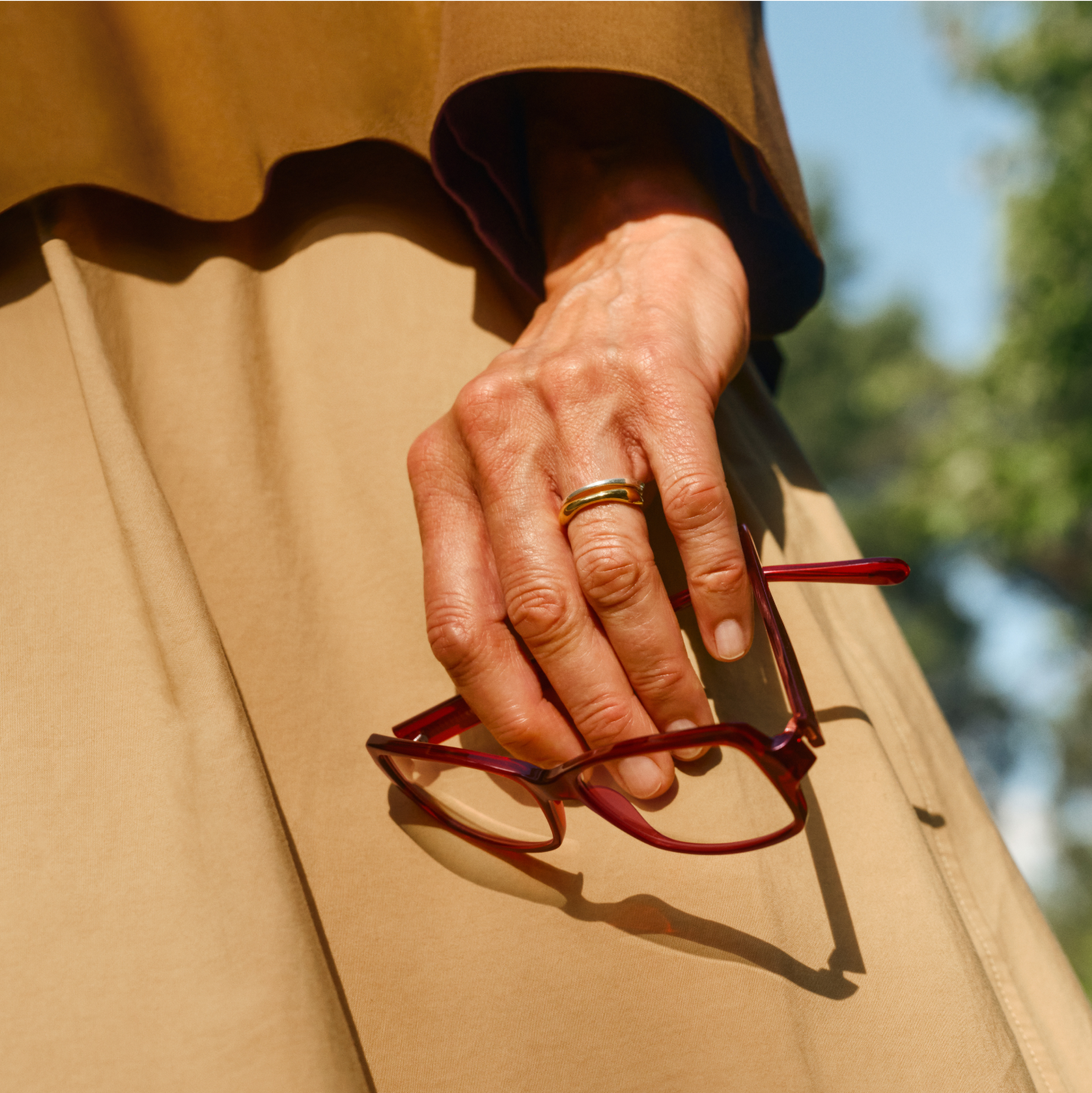 Eine Hand mit Goldring hält rote Brille vor einem beigen Rock, im Hintergrund unscharfe Außenansicht.
