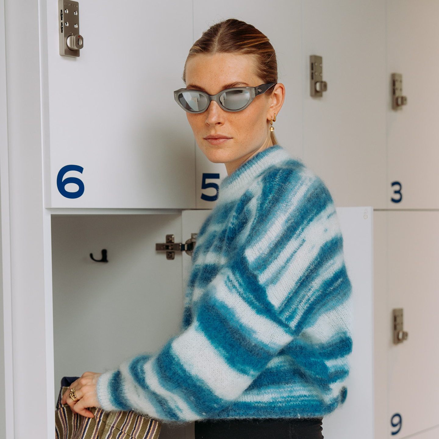 Jeune femme portant un pull rayé bleu et blanc et des lunettes de soleil, debout près des casiers, tenant un sac.