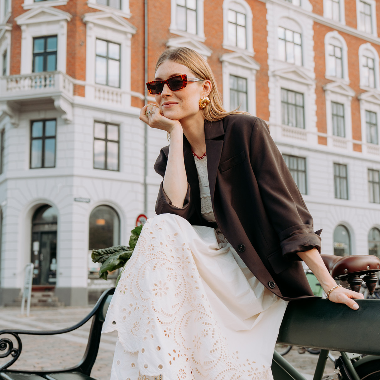 Femme avec des lunettes de soleil et un blazer sombre assise sur un banc, portant une jupe blanche ajourée, avec un bâtiment historique en arrière-plan.