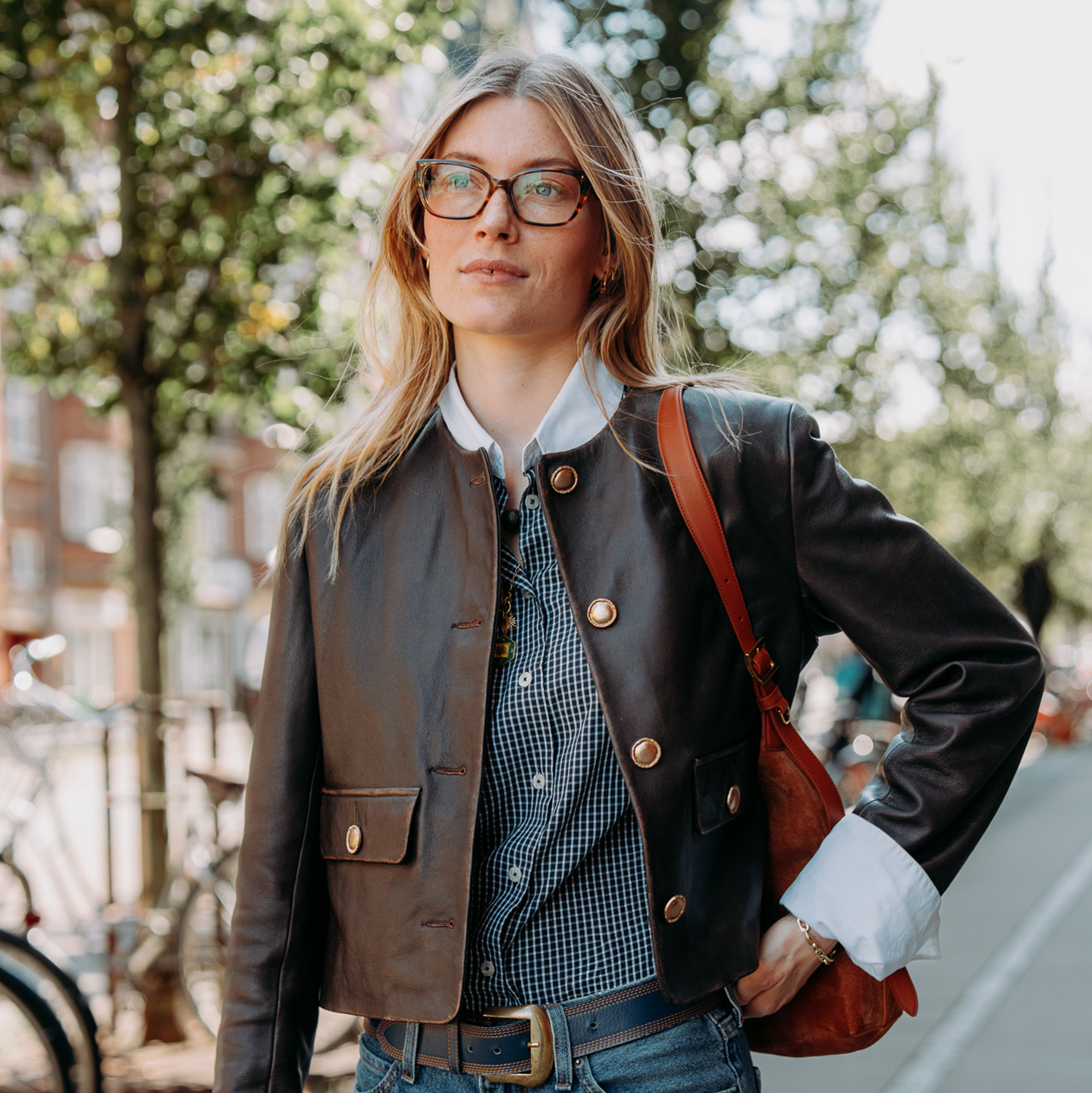 Jeune femme avec des lunettes et une veste en cuir marchant dehors, entourée d’arbres, avec une expression confiante.
