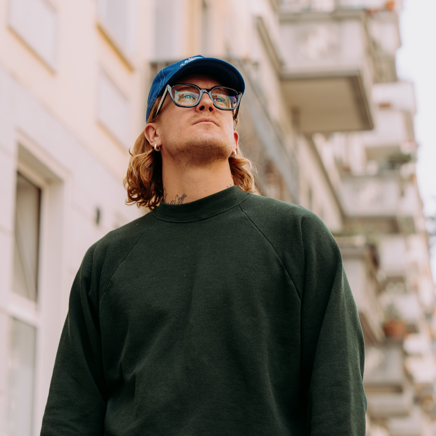 L’artiste Peter-Maximilian Ronsdorf, portant des lunettes et une casquette bleue, regarde vers le haut dans un environnement urbain.