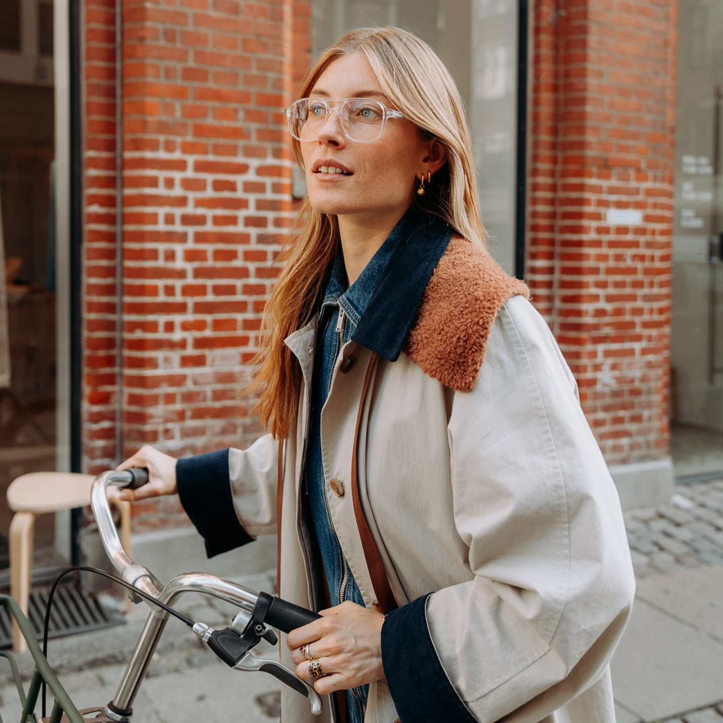 Femme aux longs cheveux et portant des lunettes, en manteau, debout avec un vélo devant un bâtiment en briques.