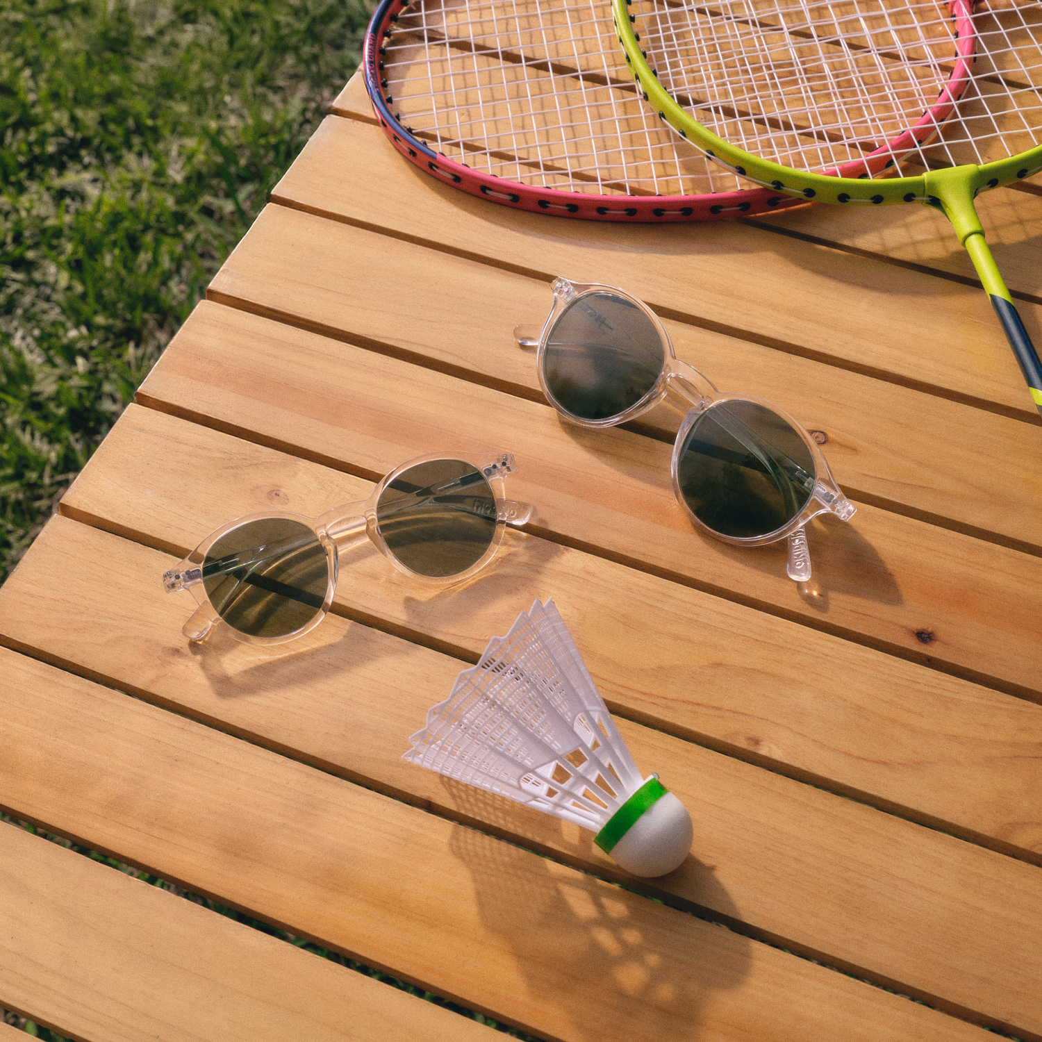 Zwei Sonnenbrillen liegen auf einem Holztisch neben Badmintonschlägern und einem Federball.