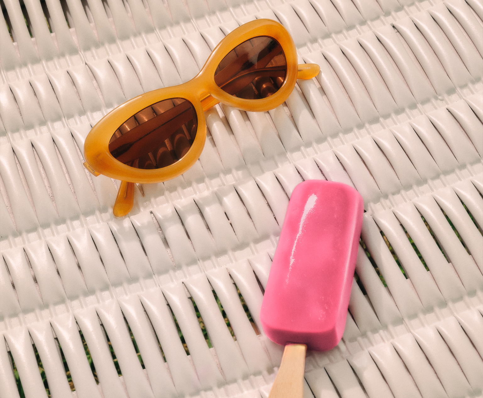 Eine gelbe Cat-Eye-Sonnenbrille und ein rosa Eis am Stiel auf einer weißen Korbfläche.