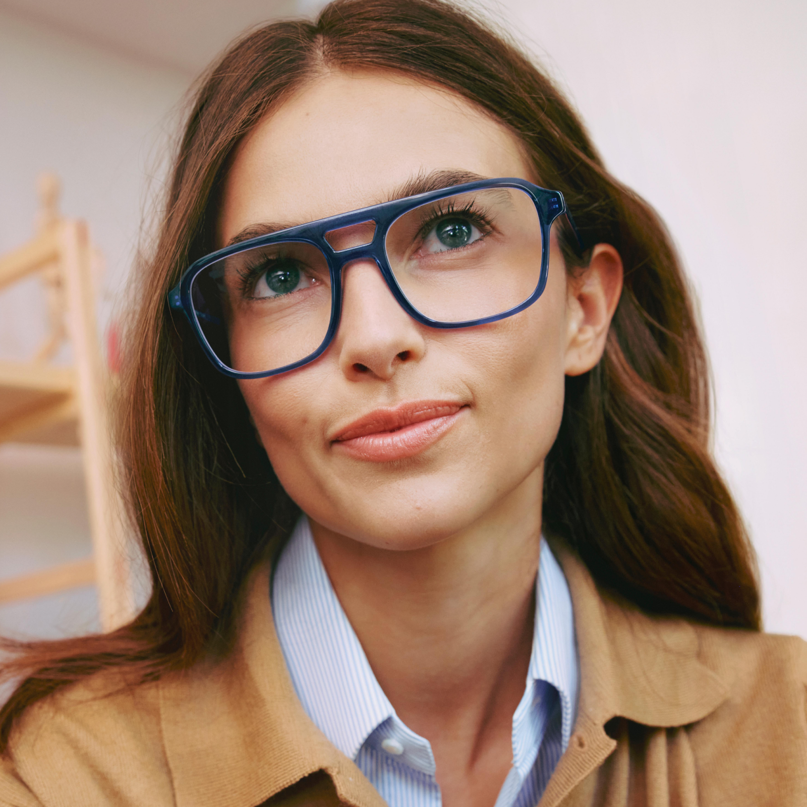 Eine Frau mit langen braunen Haaren trägt eine große Brille mit blauem Rahmen und einen beigefarbenen Pullover über einem gestreiften Oberteil und blickt zur Seite.