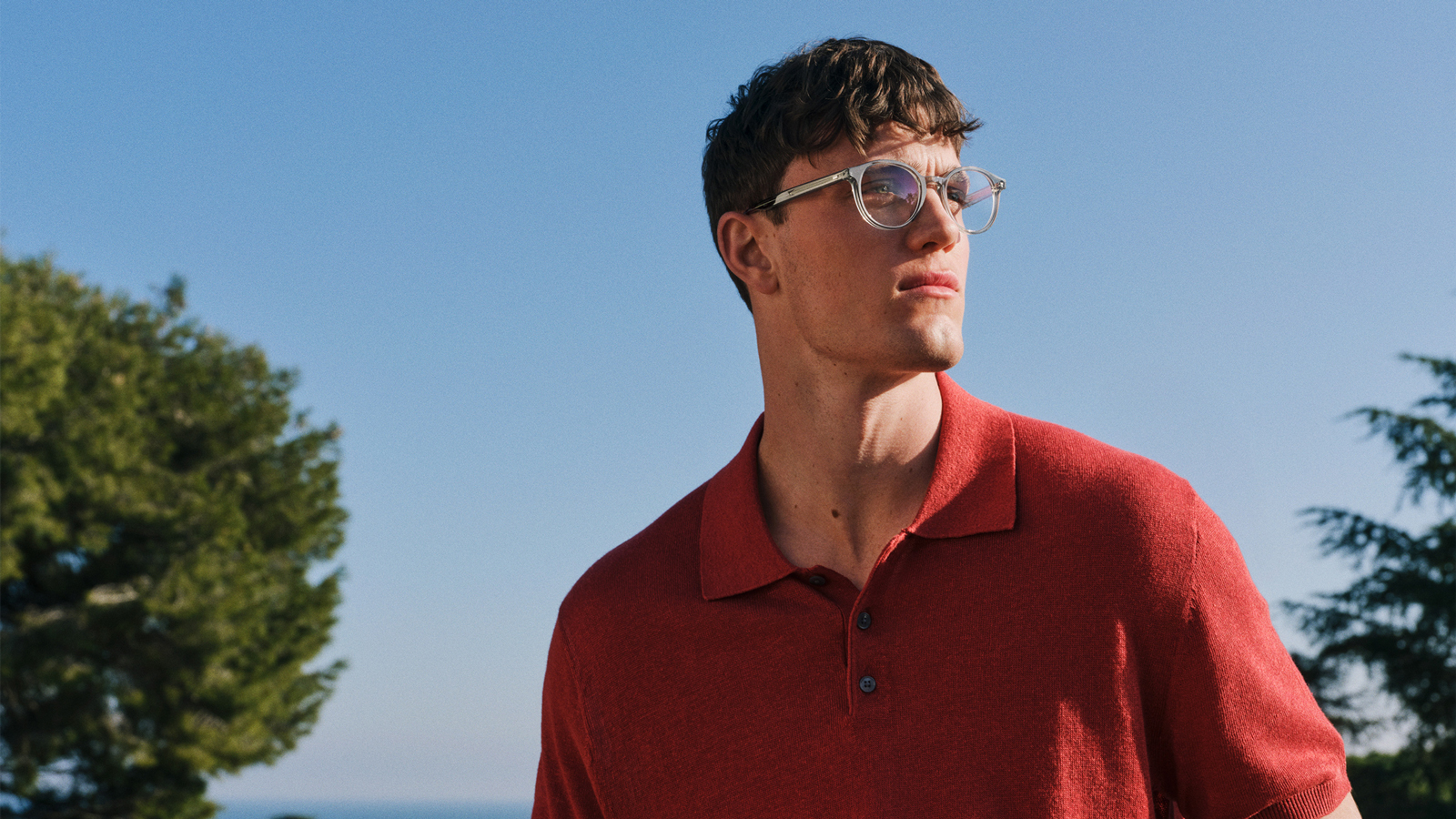 Ein Mann in einem roten Poloshirt und einer klaren Brille blickt zur Seite, im Hintergrund sind Bäume und der blaue Himmel zu sehen.