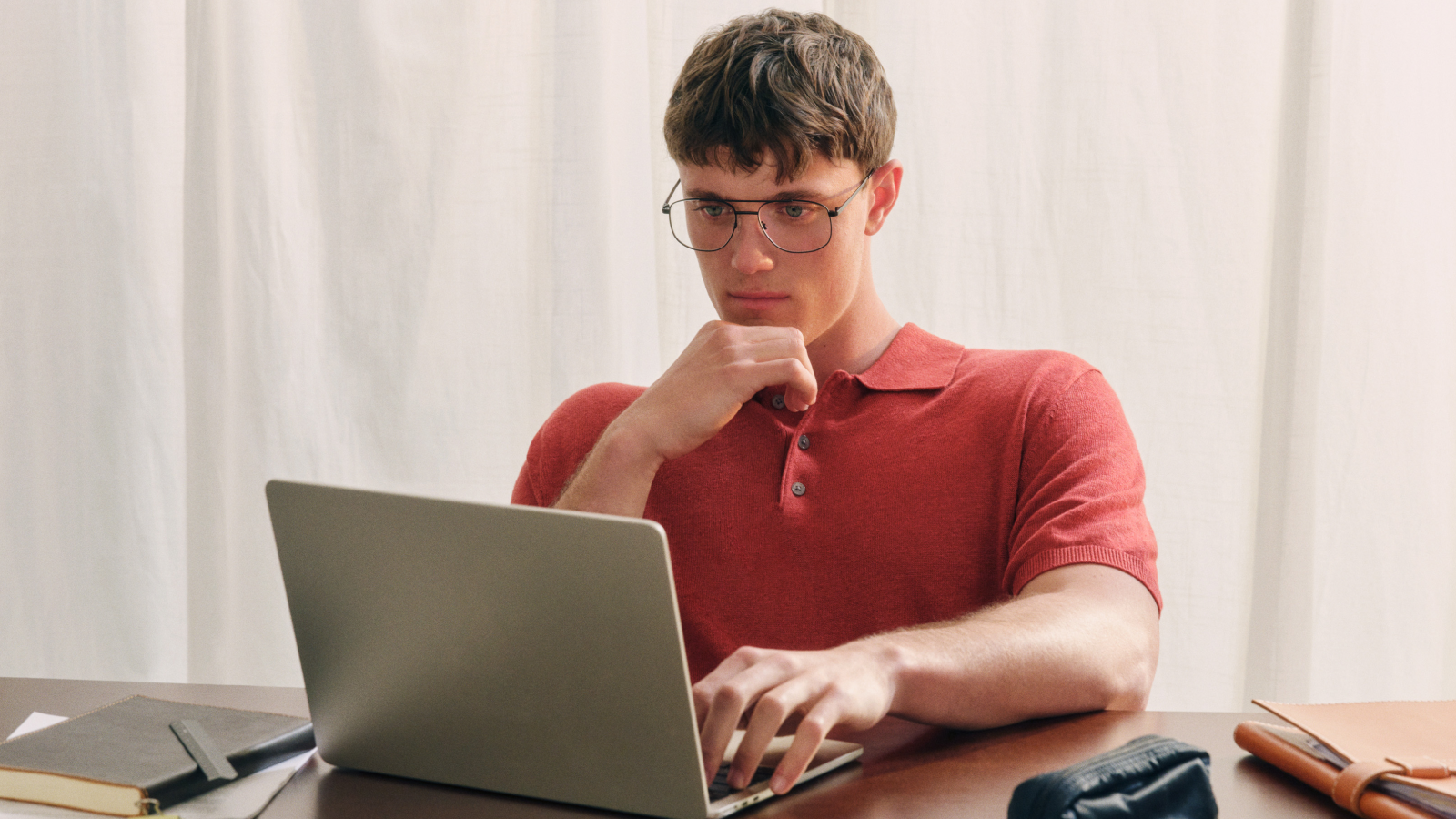 Person trägt eine Brille und ein rotes Hemd und blickt nachdenklich auf den Bildschirm eines Laptops. Die Person sitzt vor einem hellen Vorhang.