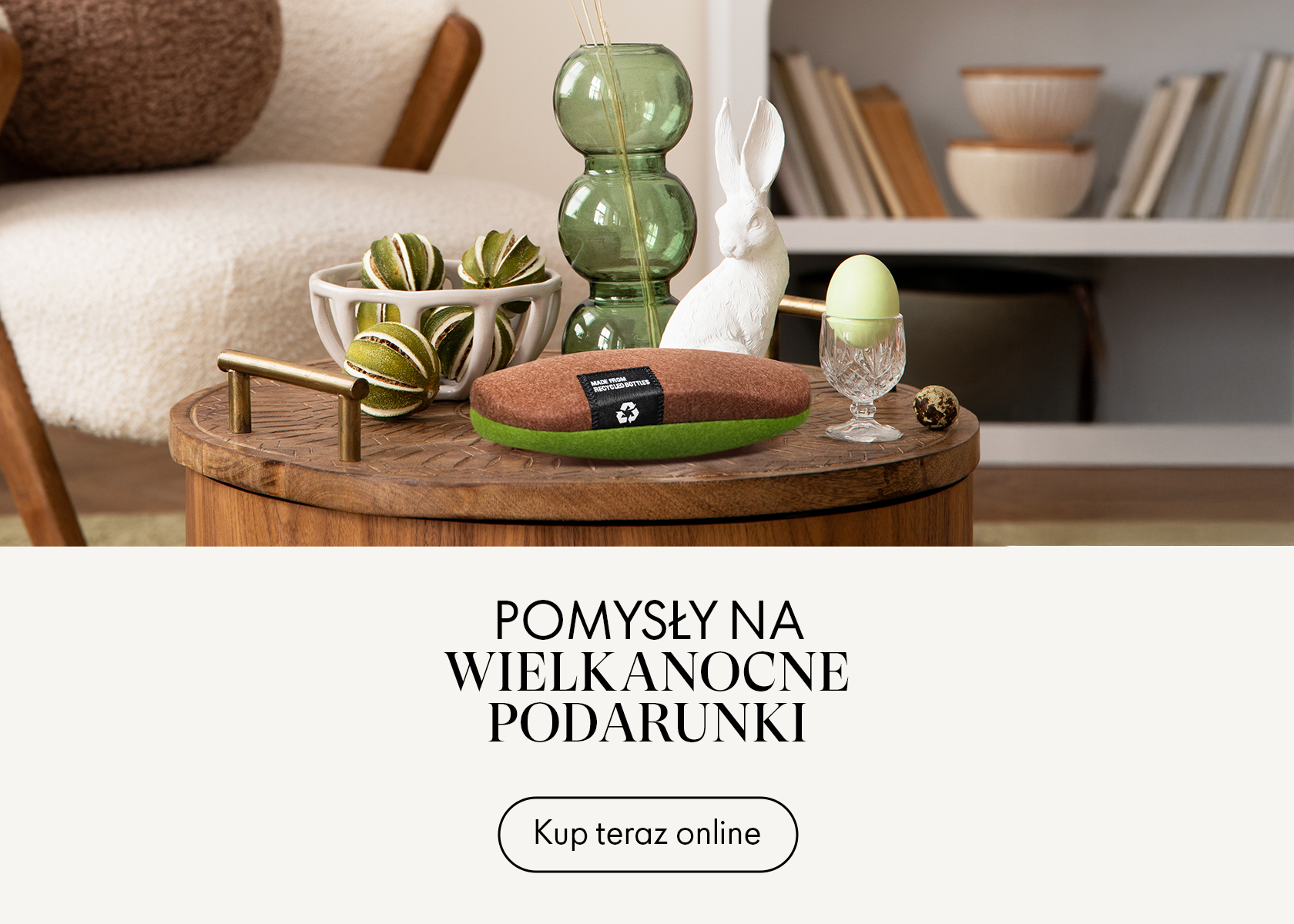 Przytulny salon z drewnianym stołem, wielkanocnymi dekoracjami, figurką białego królika, zielonymi wazonami oraz tekstem reklamowym z pomysłami na wielkanocne prezenty.