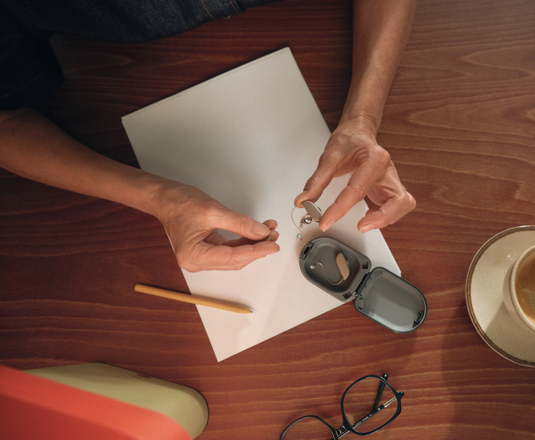 Eine Person wechselt die Batterie eines Hörgeräts an einem Holztisch, auf dem Papier, ein Bleistift, eine Brille und eine Tasse Kaffee liegen.