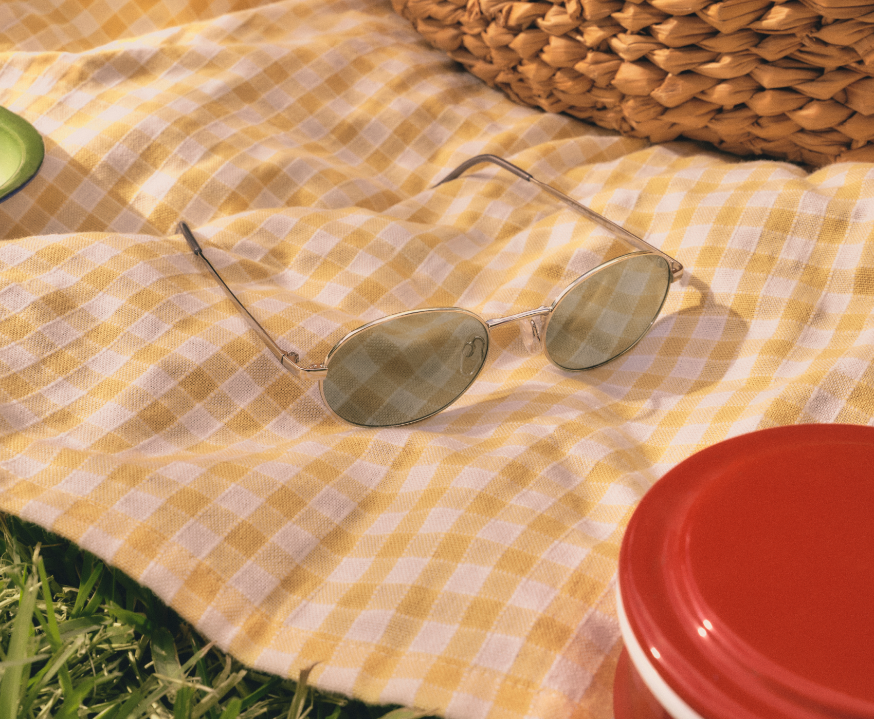 Sonnenbrille auf einer gelb karierten Picknickdecke, neben einem geflochtenen Korb und einem roten Behälter, aus dem Gras herausschaut.