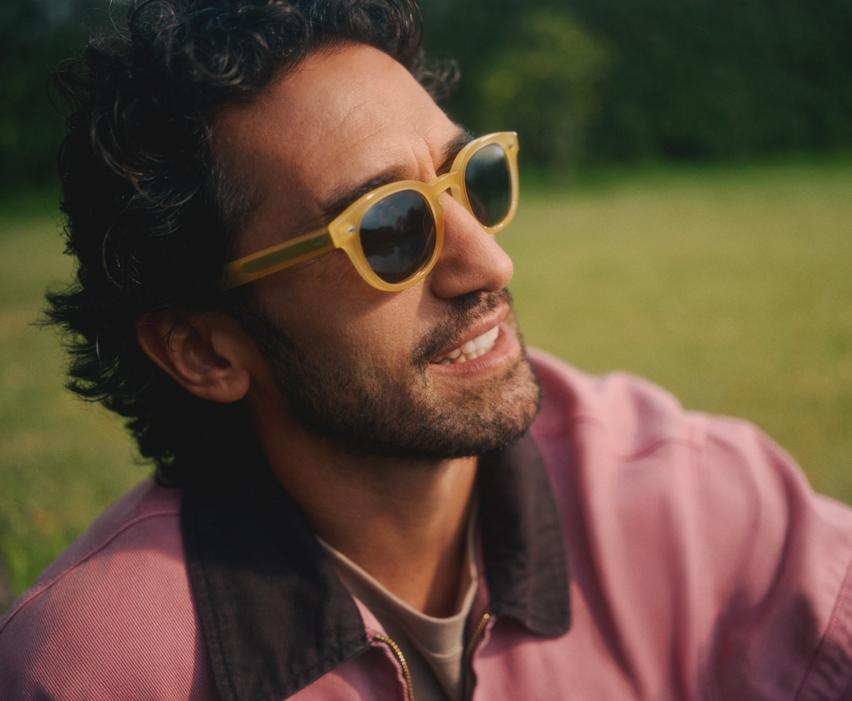 Un homme portant des lunettes de soleil et une veste violette est assis à l'extérieur, le regard pensif tourné vers le ciel, avec de l'herbe verte en arrière-plan.