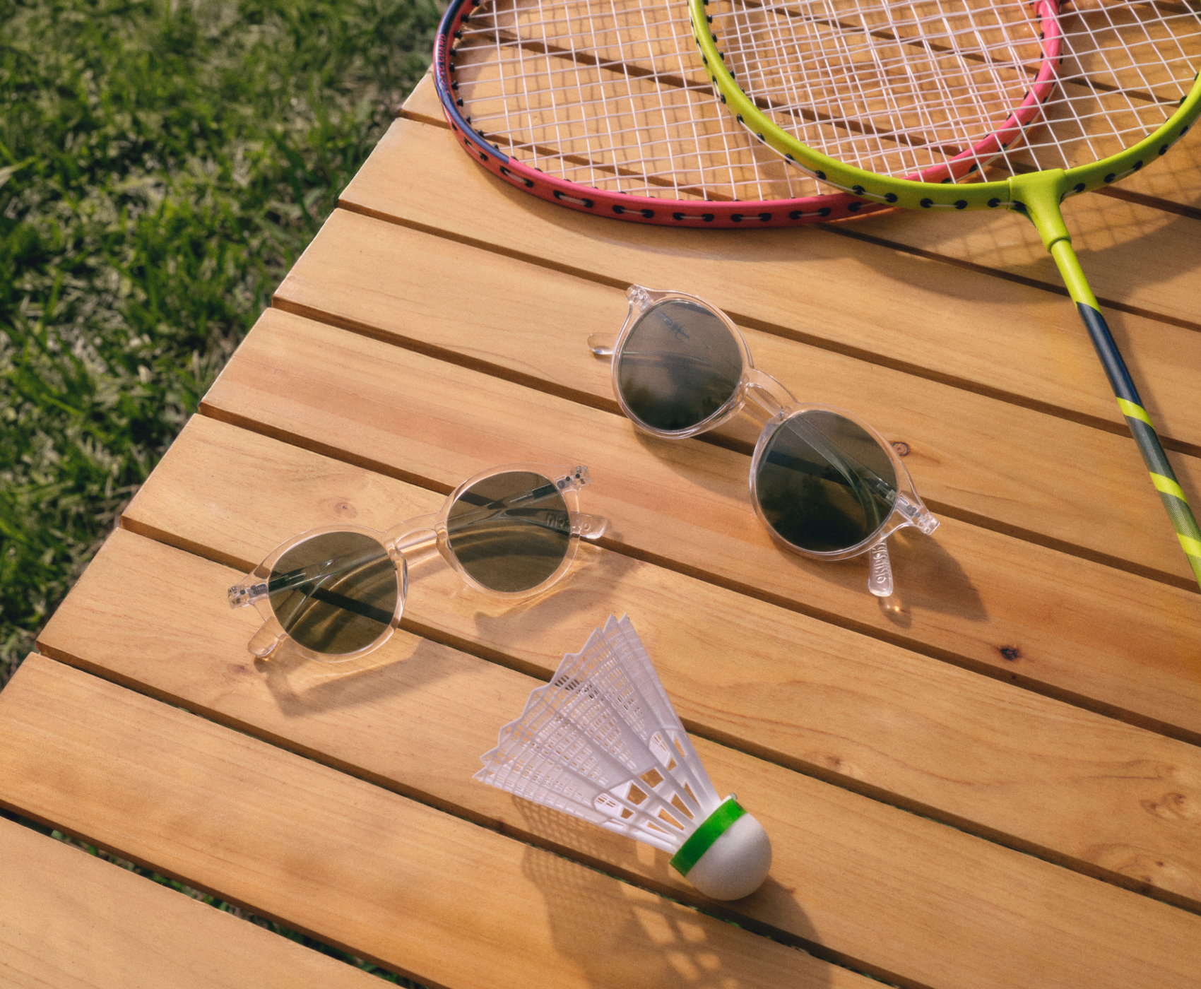 Sonnenbrille und Federball auf einem Holztisch neben Badmintonschlägern, auf einer Rasenfläche.