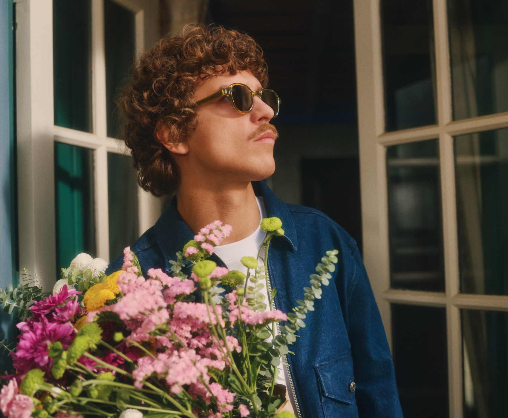 Personne aux cheveux bouclés et portant des lunettes de soleil, tenant un bouquet de fleurs colorées, debout près de grandes fenêtres et vêtue d'une veste bleue.