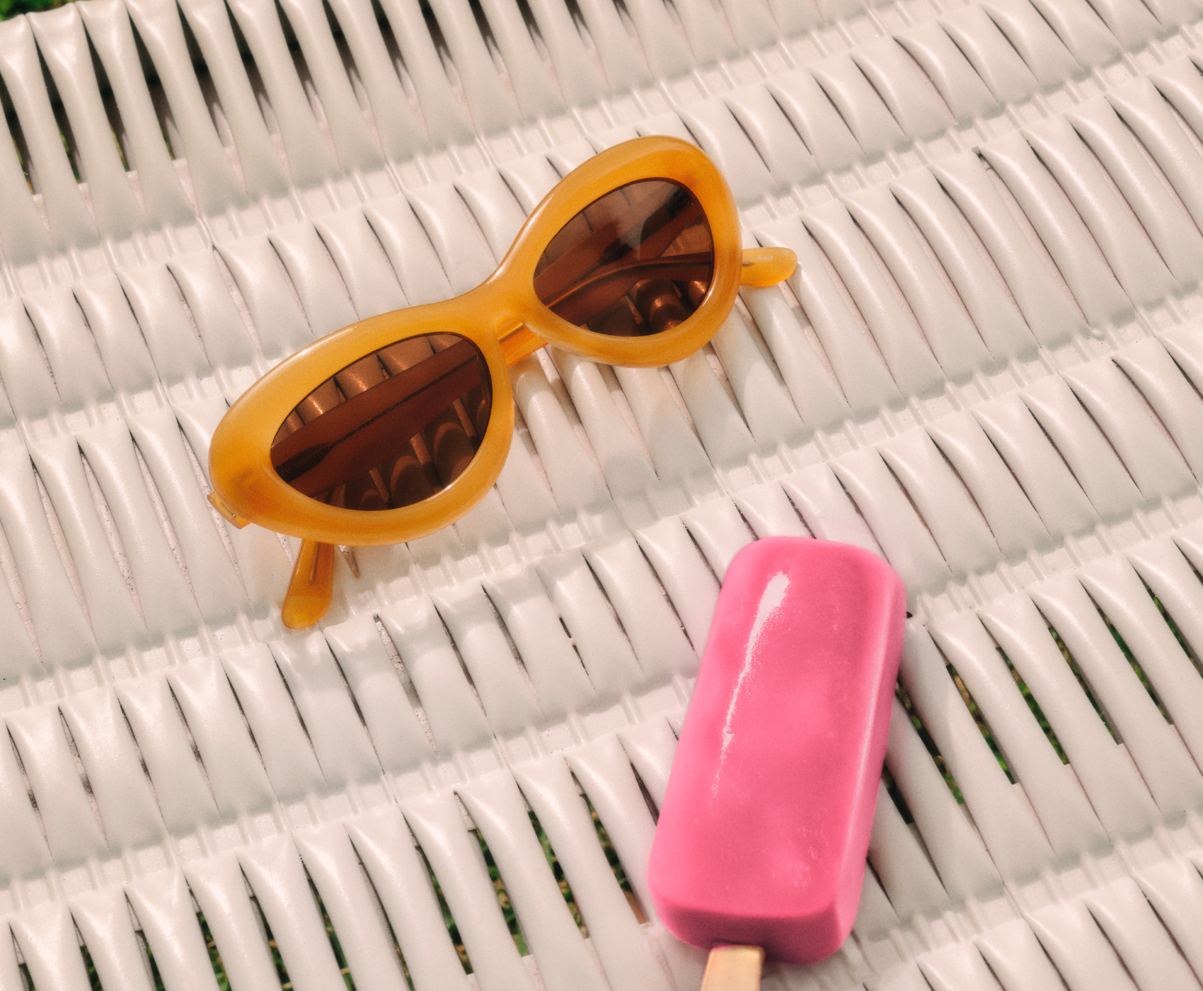 Gelbe Sonnenbrille und ein rosa Eis am Stiel liegen auf einer weißen Weidenoberfläche, im Hintergrund grünes Gras.