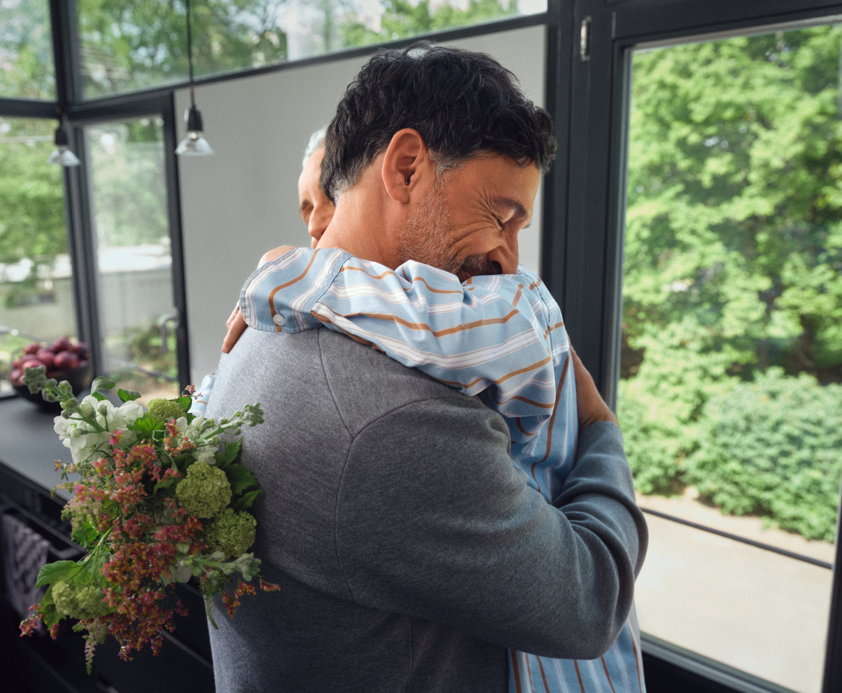Ein Mann in einem grauen Pullover umarmt eine Person in einem gestreiften Hemd und lächelt herzlich, während im Hintergrund ein Blumenstrauß zu sehen ist.