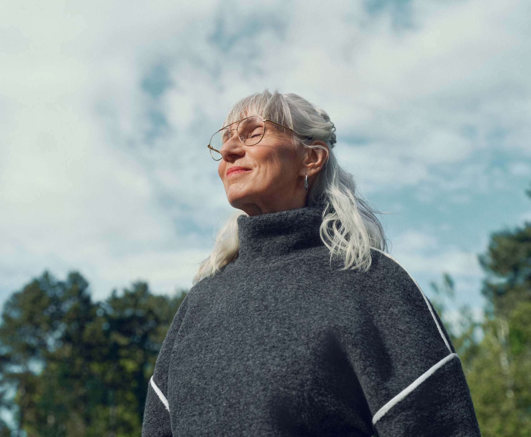 Une femme âgée aux longs cheveux blancs et portant des lunettes, vêtue d’un pull gris, se tient dehors sous un ciel nuageux, l’air serein.