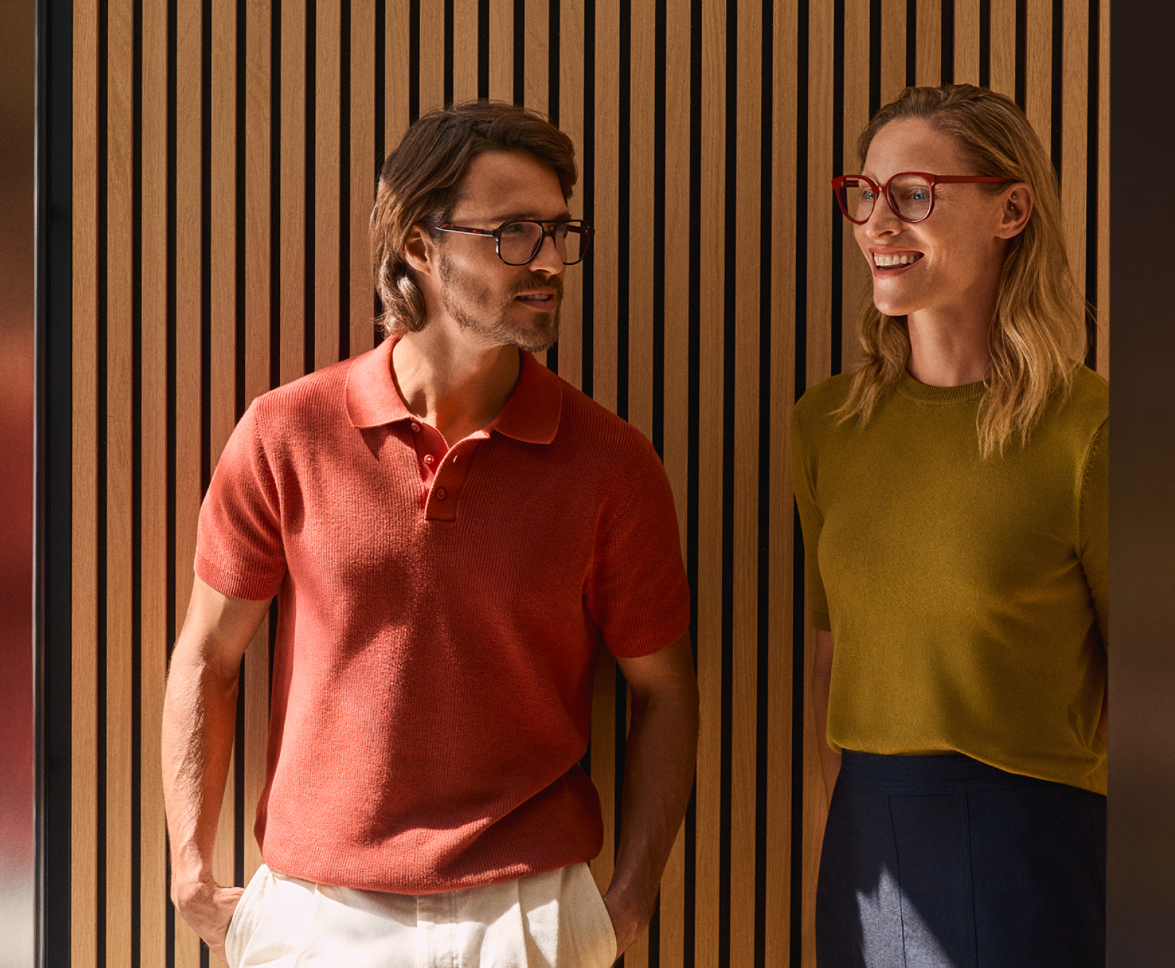 Un uomo con un polo rosso e una donna con una camicia verde, entrambi con gli occhiali, stanno sorridenti davanti a una parete di listelli di legno.