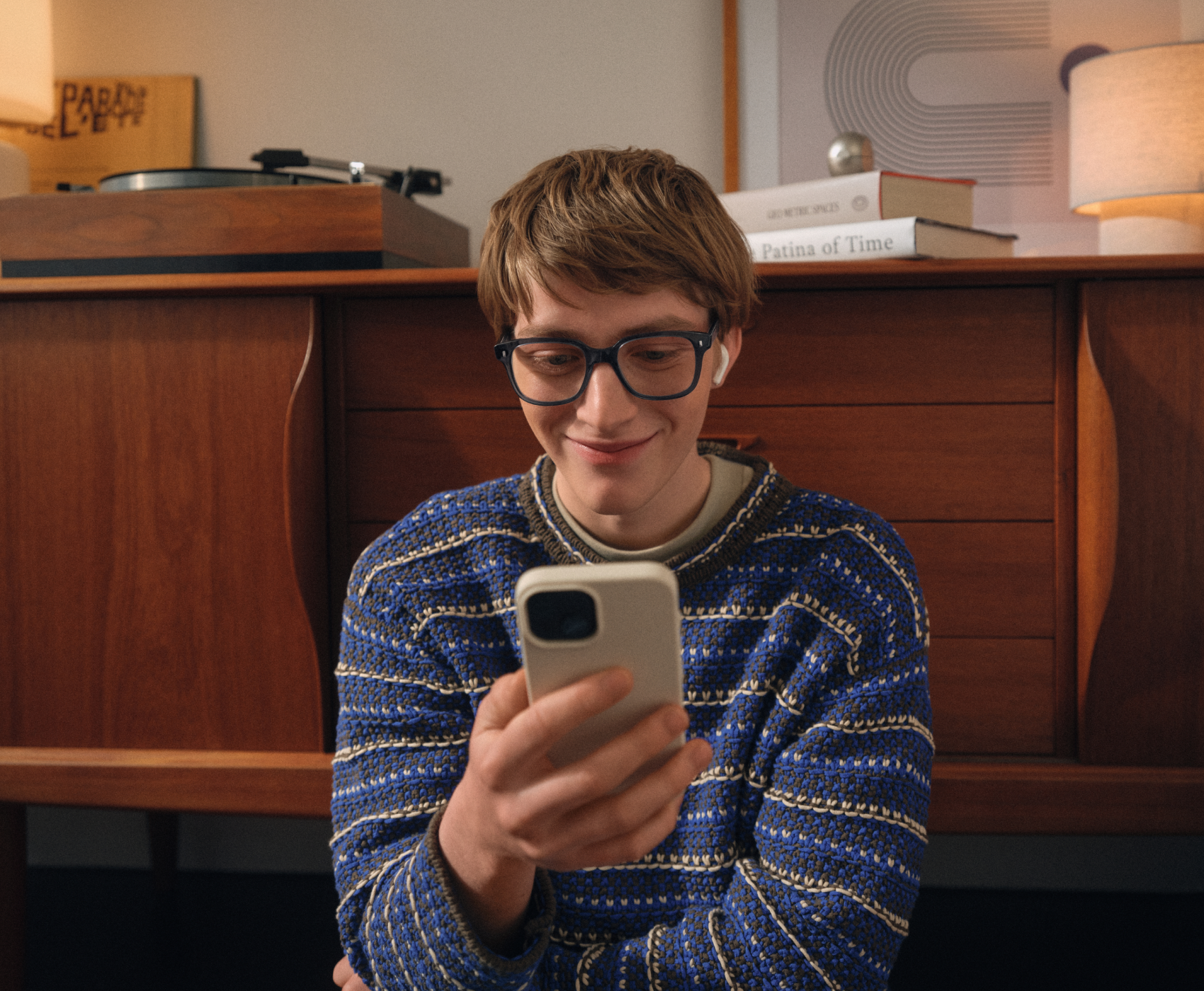 Una persona con gli occhiali e un maglione a righe sorride mentre guarda uno smartphone. Sullo sfondo ci sono un armadio di legno e dei libri.