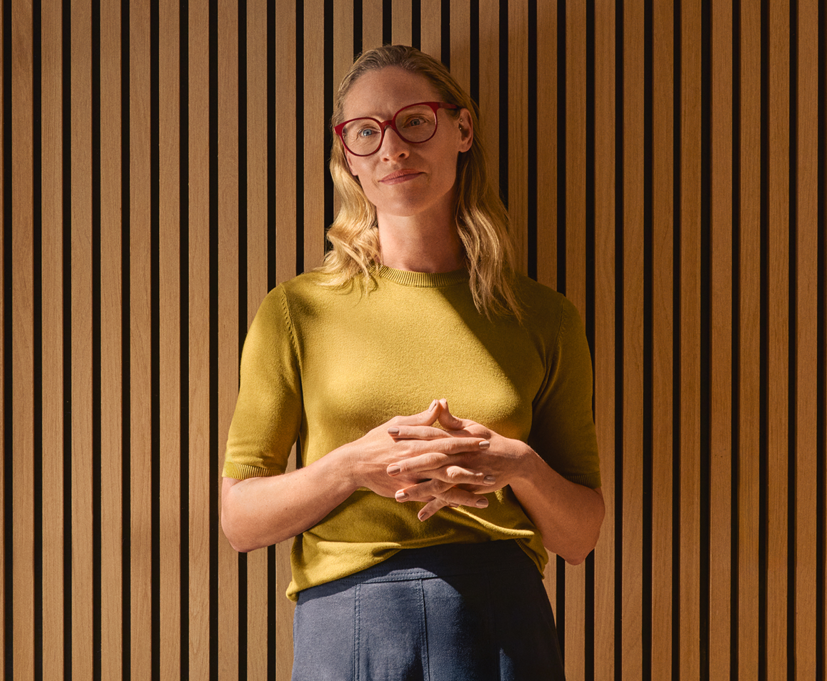 Donna con capelli biondi e occhiali rossi si trova contro una parete di listelli di legno.