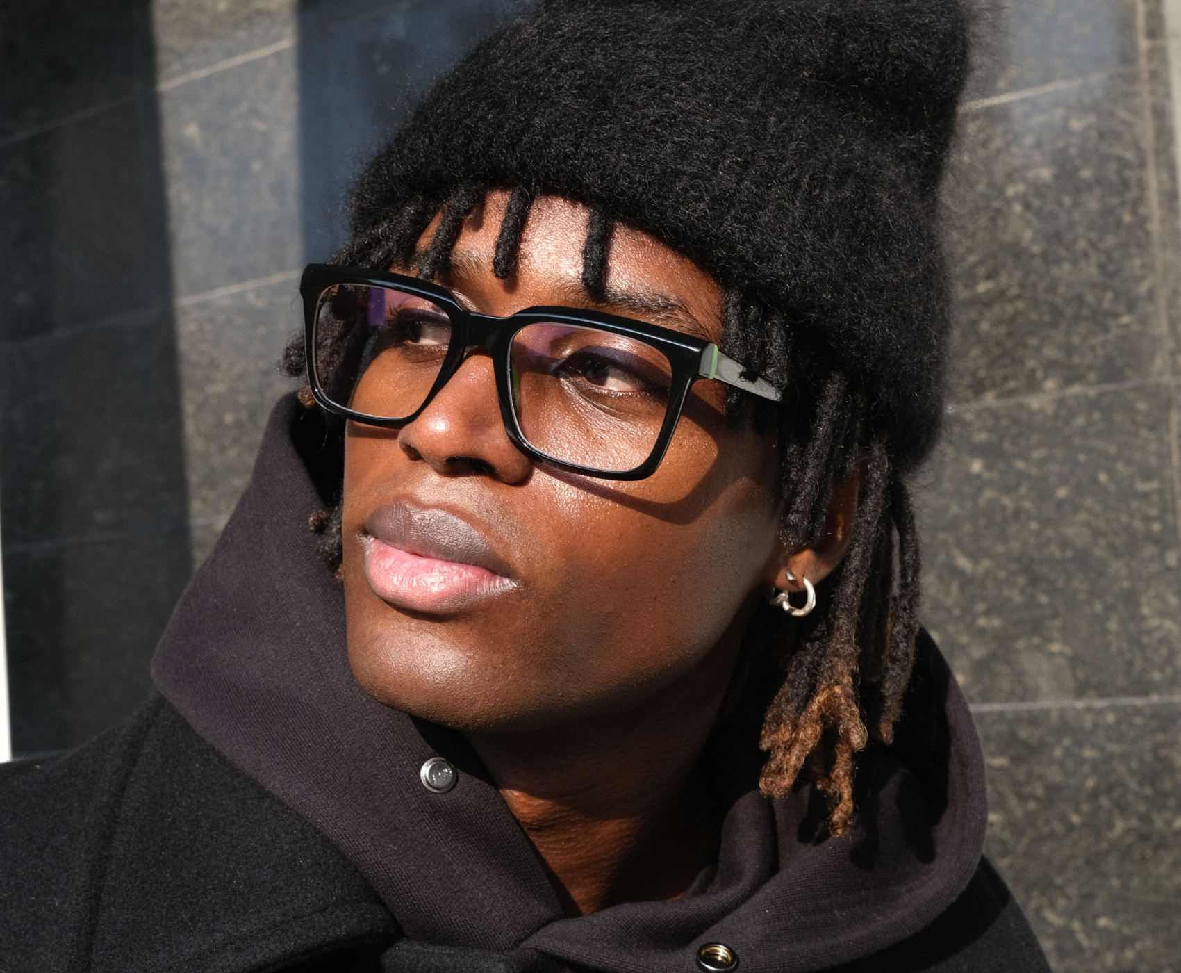 Uomo con berretto nero, occhiali e cappotto scuro guarda di lato, mentre la luce del sole proietta ombre su un muro di pietra dietro di lui.