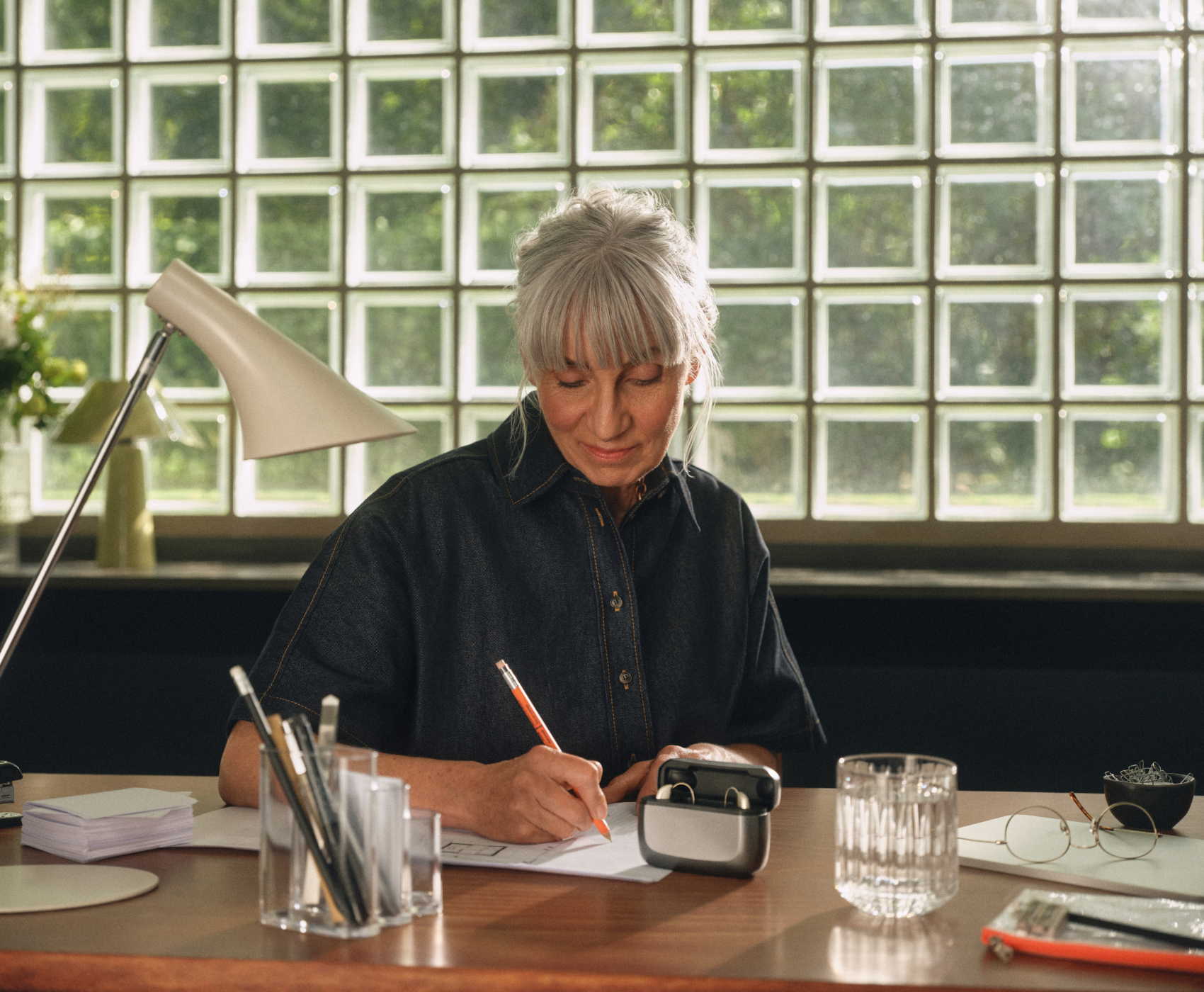 Ältere Frau mit grauen Haaren schreibt an einem Schreibtisch mit einem Bleistift. Eine Lampe und ein Behälter mit Stiften stehen in der Nähe, im Hintergrund sind Glasbausteinfenster zu sehen.