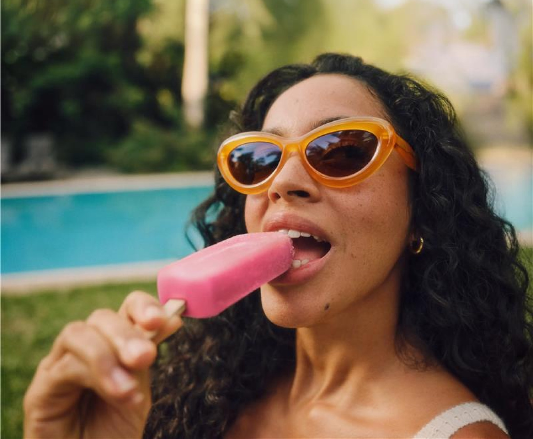 Frau mit orangefarbener Sonnenbrille, die draußen ein rosa Eis am Stiel genießt. Lockiges Haar, große Creolen, sonniger Hintergrund.