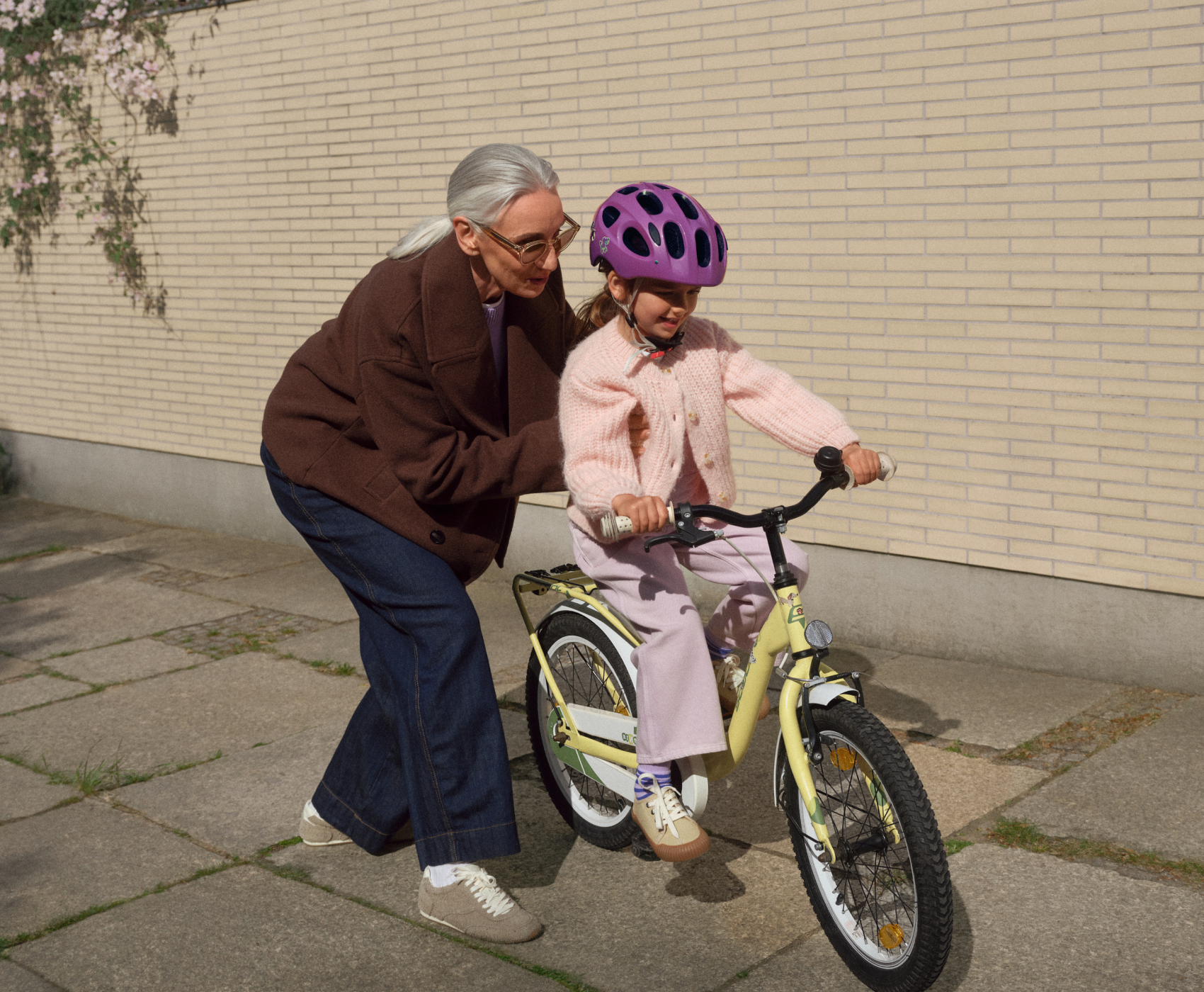 Eine ältere Frau hilft einem jungen Mädchen mit einem rosa Helm, auf einem gepflasterten Weg in der Nähe einer Backsteinmauer mit einem gelben Fahrrad zu fahren.