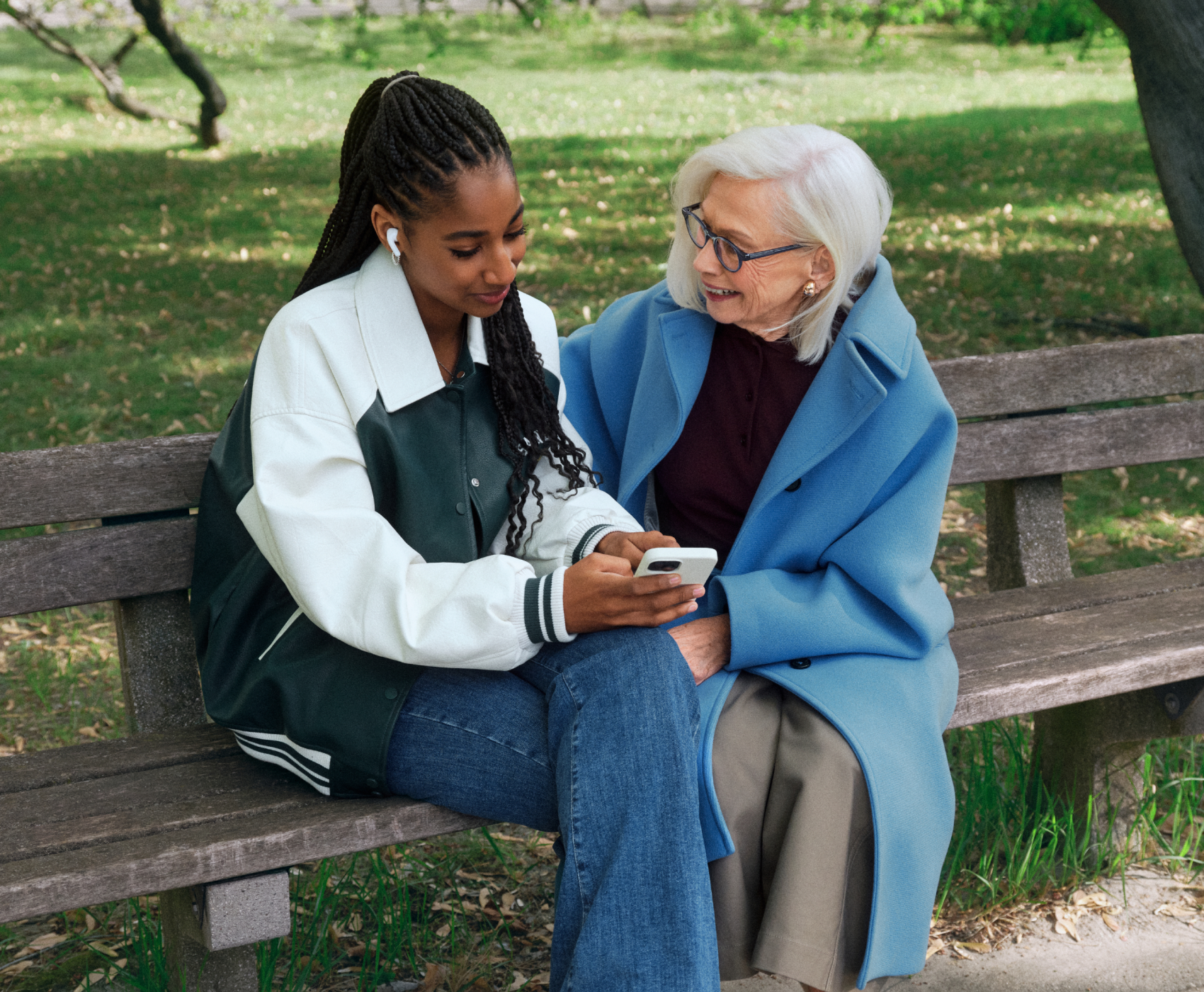 Zwei Frauen sitzen auf einer Parkbank – eine junge mit geflochtenen Haaren und Ohrhörern, die andere älter mit weißen Haaren und Brille. Beide schauen auf ein Smartphone.