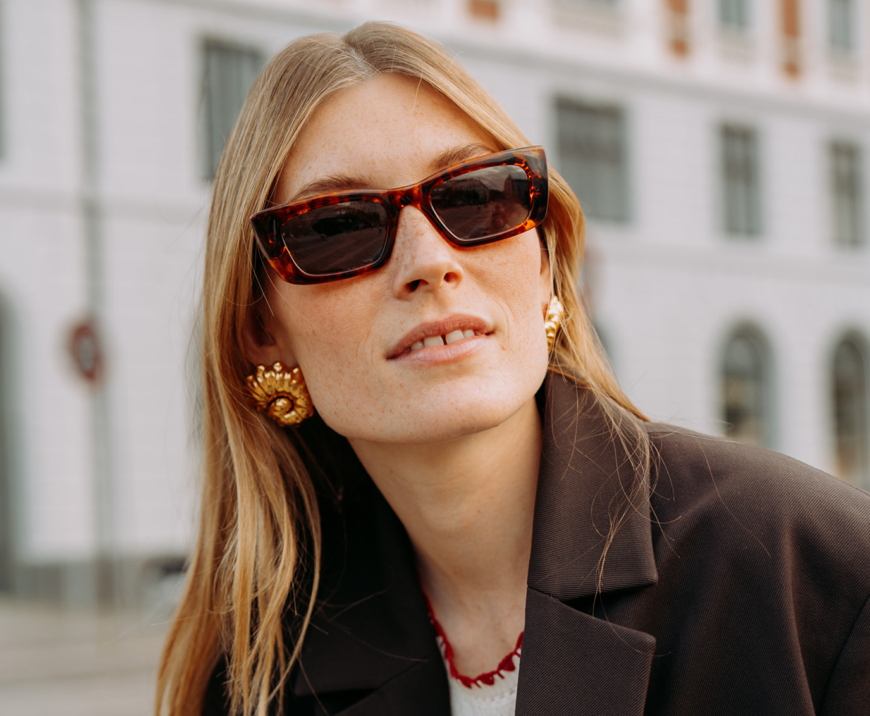 Eine Frau mit langen Haaren trägt havannafarbene Sonnenbrillen, große goldene Ohrringe und einen Blazer. Sie steht im Freien und blickt in die Ferne.