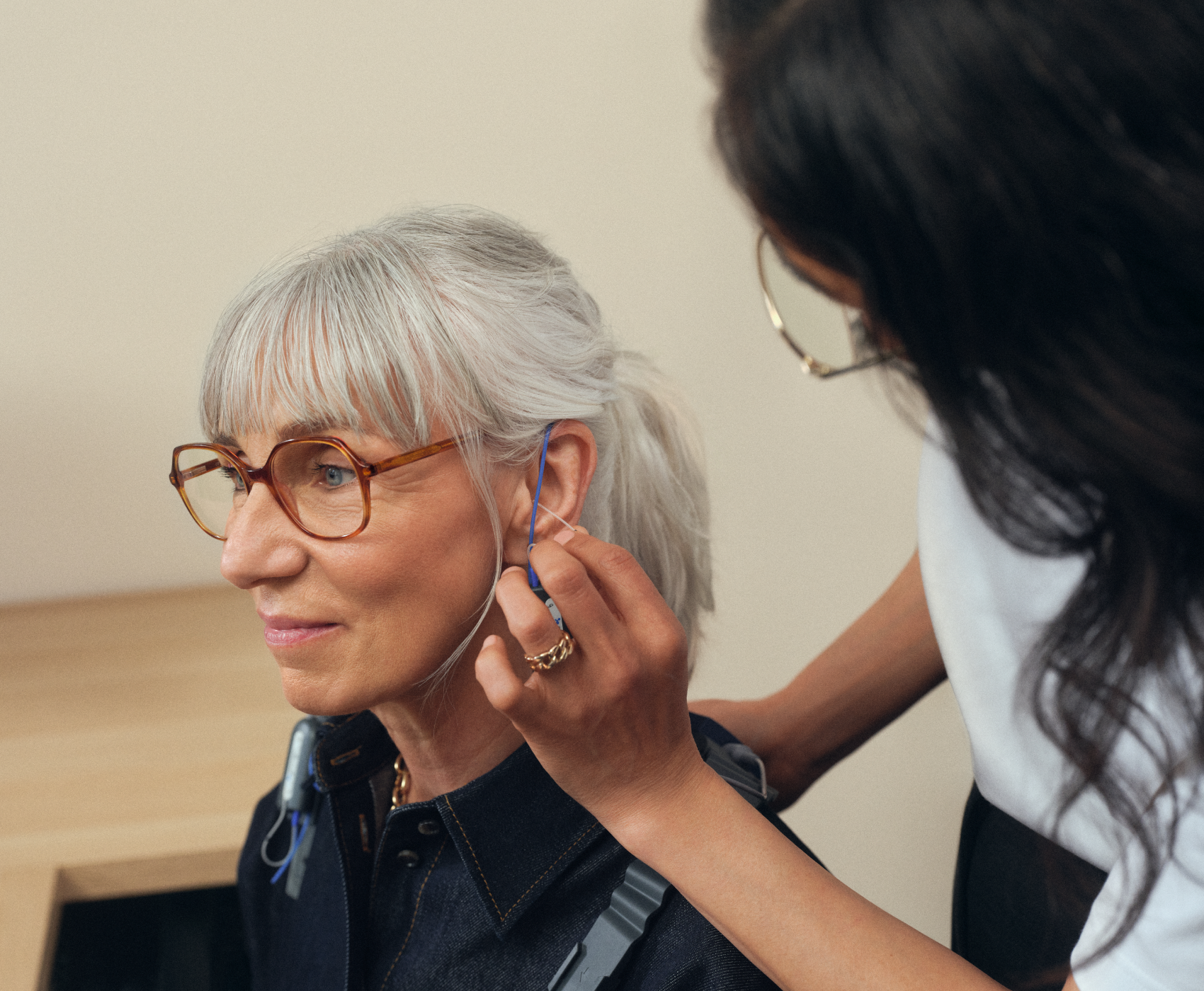 Une femme aux cheveux gris et portant des lunettes se fait poser un appareil auditif par une autre personne dans une pièce bien éclairée.
