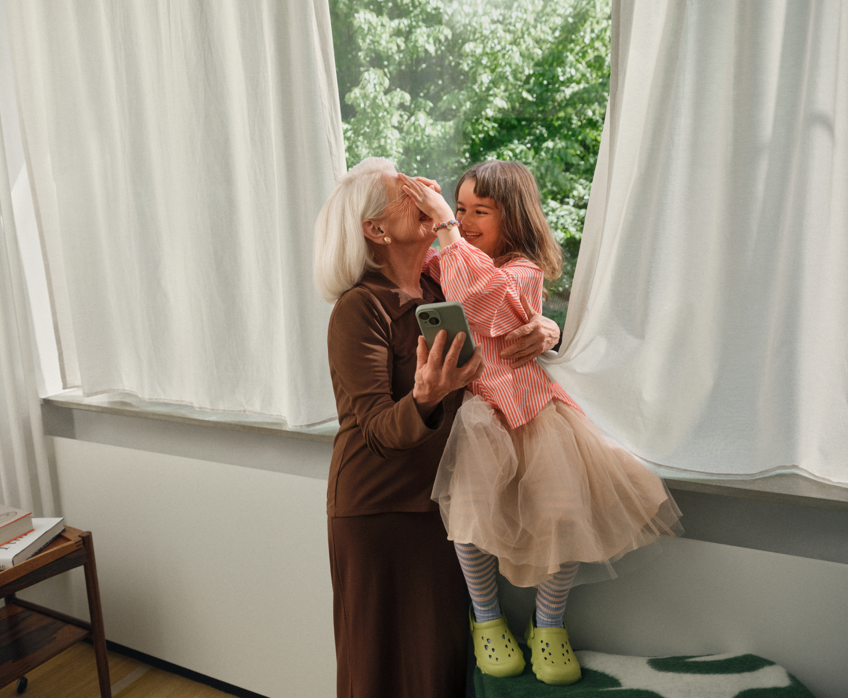 Ältere Frau und junges Mädchen lachen in der Nähe eines Fensters; die Frau hält ein Telefon, und das Mädchen bedeckt spielerisch ihre Augen.