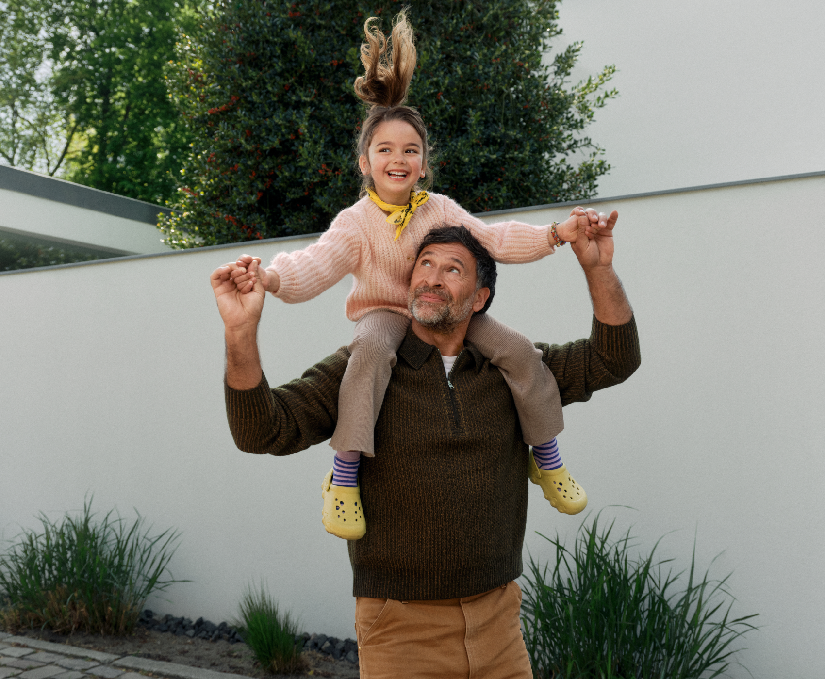 Un homme porte un enfant souriant sur son dos dans un jardin. Tous deux se tiennent par la main et regardent vers le haut.