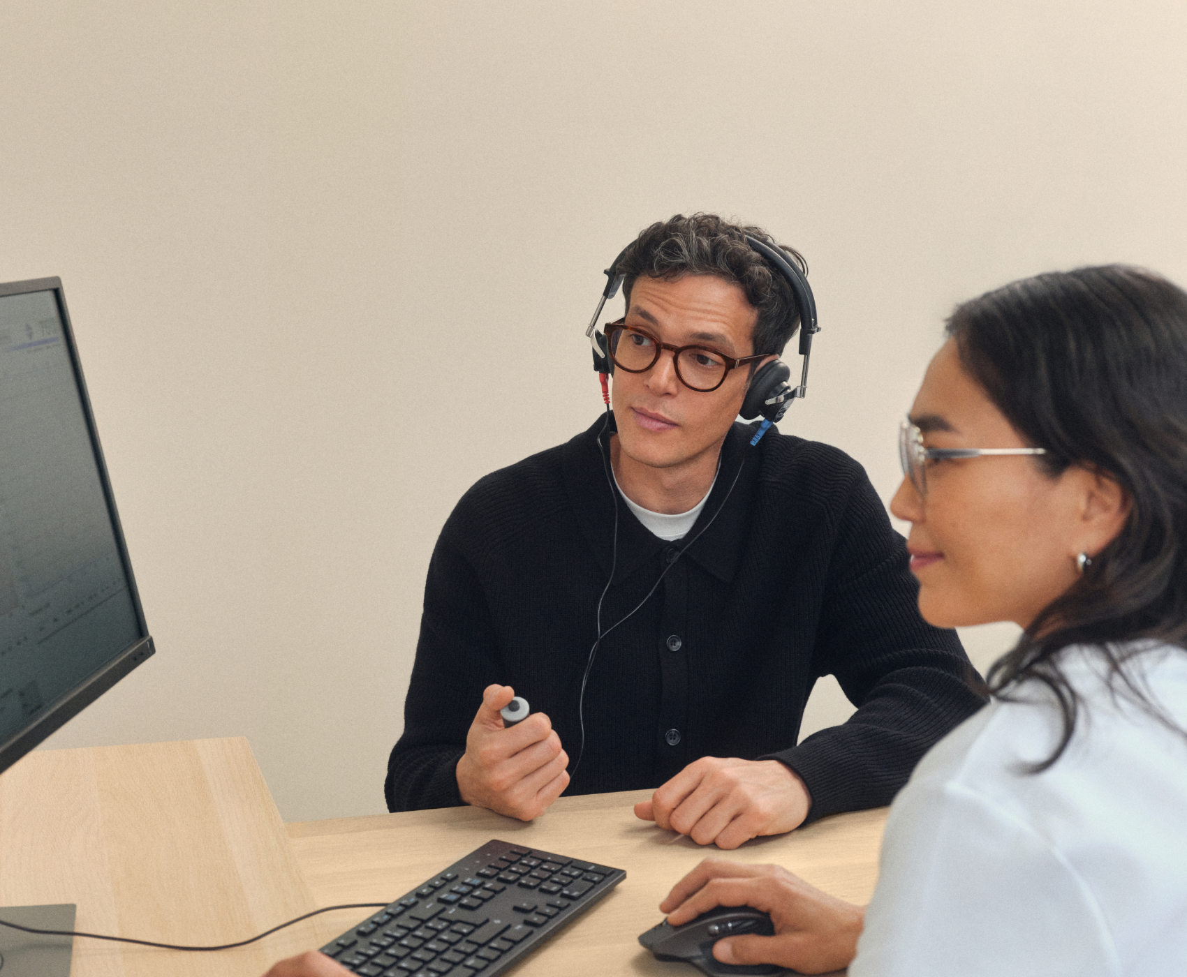 Zwei Personen an einem Schreibtisch, eine davon trägt Kopfhörer und hält einen Stift in der Hand, beide konzentrieren sich auf einen Computerbildschirm.