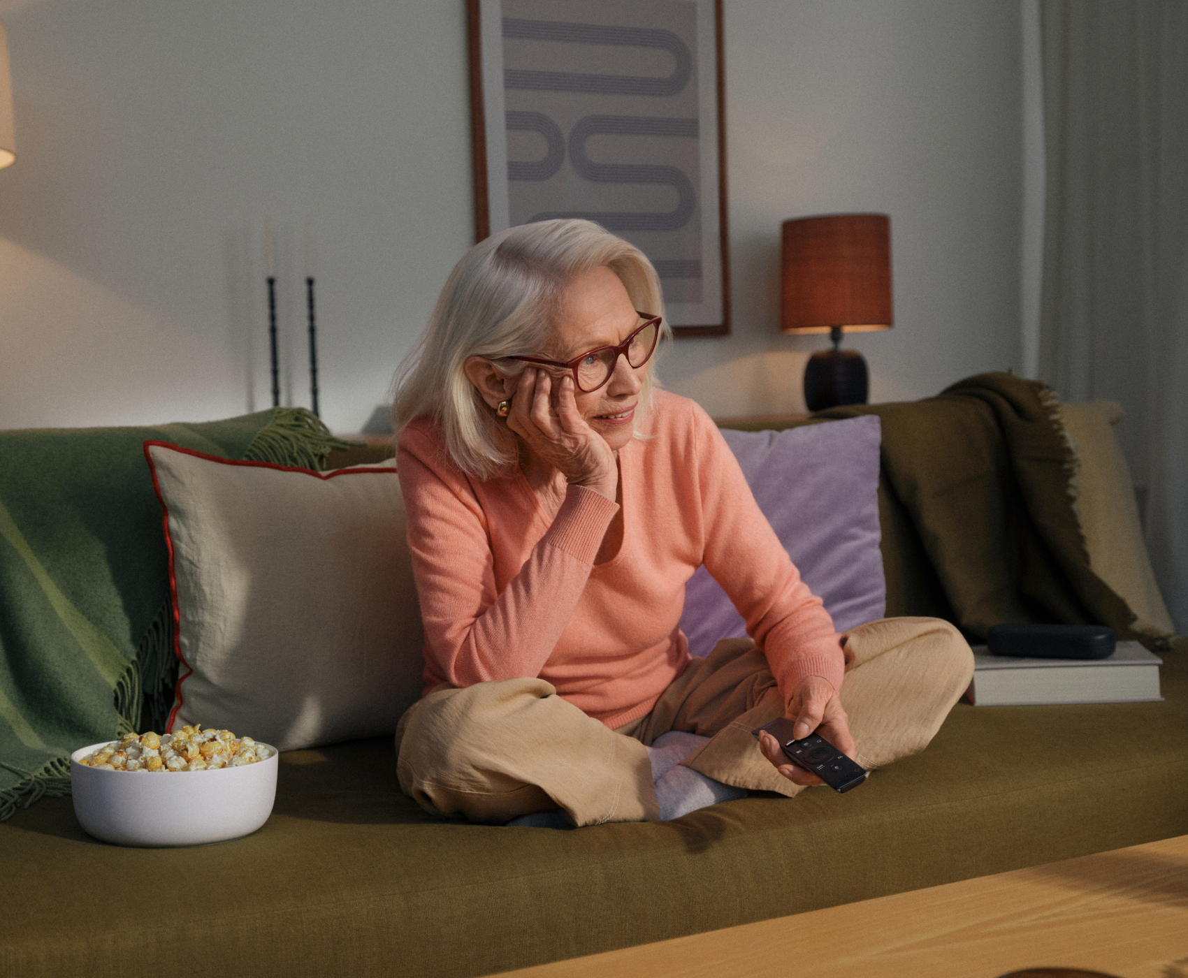 Eine ältere Frau mit Brille sitzt auf einer Couch und hält eine Fernbedienung in der Hand.