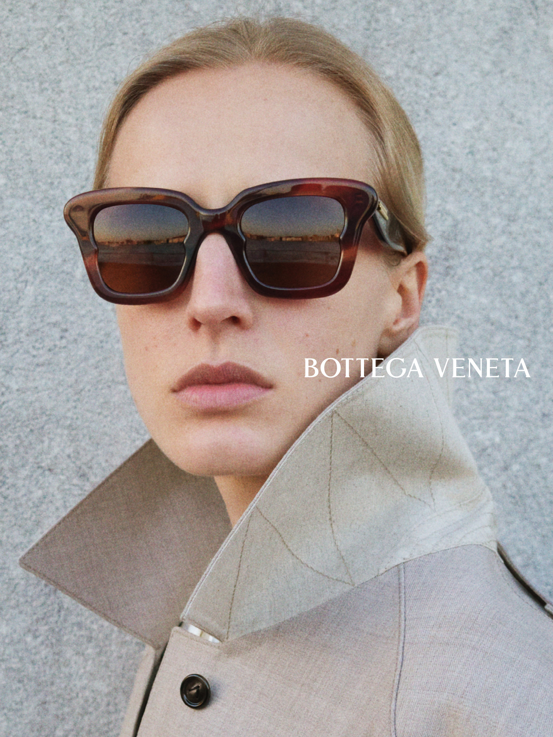 Une personne portant de grandes lunettes de soleil foncées et un manteau clair au col relevé, sur le côté duquel figure l'inscription « Bottega Veneta » sur fond gris.