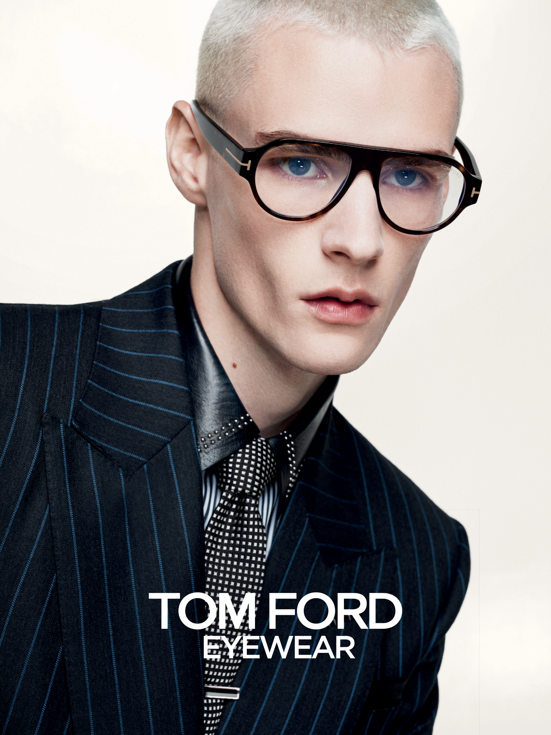 Eine Person mit kurzen Haaren trägt eine stilvolle Brille und einen Nadelstreifenanzug und posiert vor einem hellen Hintergrund für Tom Ford Eyewear.