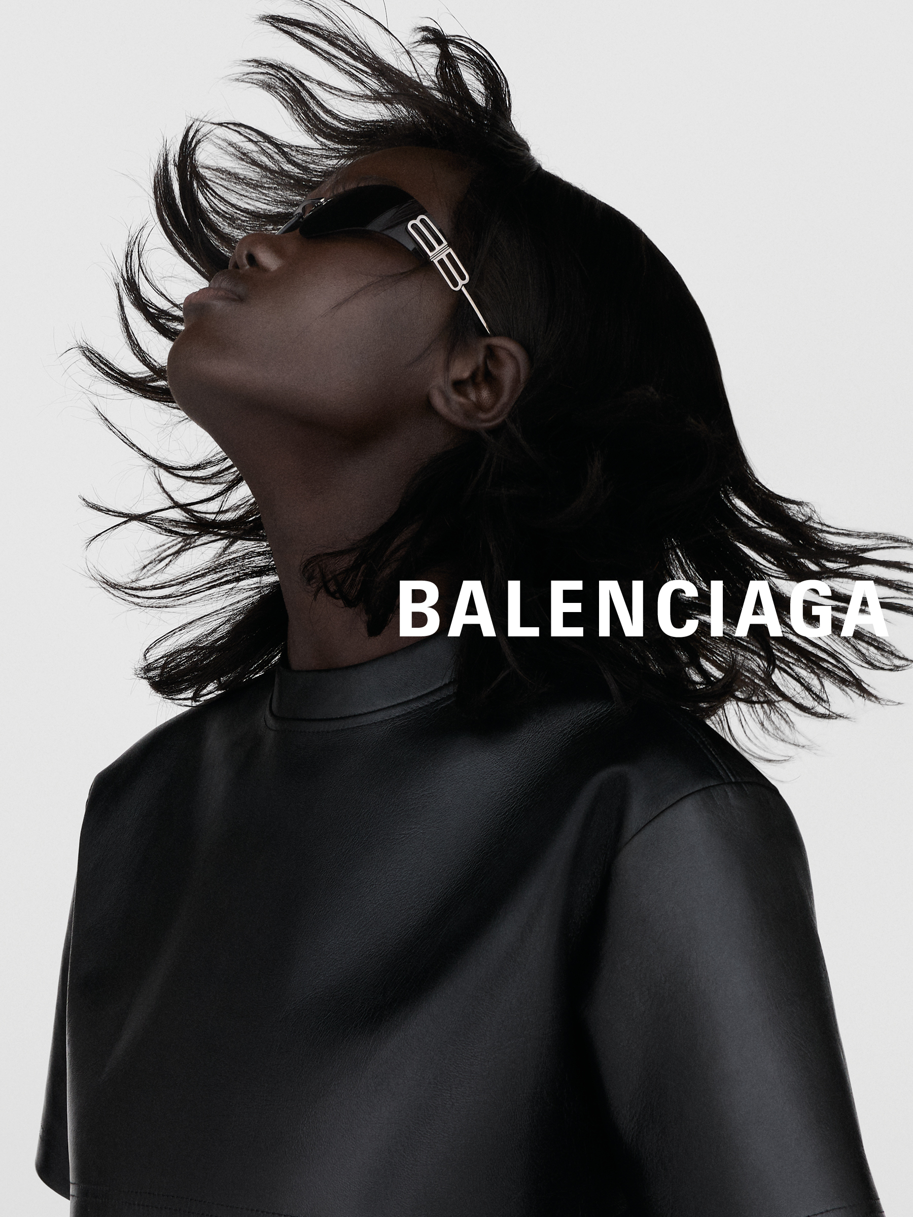 Person in schwarzem Lederoutfit und Sonnenbrille neigt den Kopf nach hinten, wobei ihr Haar herabfließt, mit dem Schriftzug „BALENCIAGA” über dem Bild.