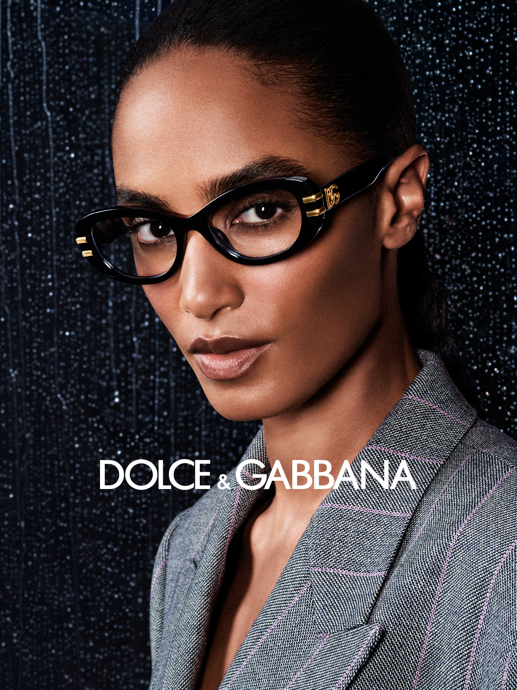 Su uno sfondo scuro e strutturato si vede una persona che indossa occhiali e un completo grigio. È raffigurato il logo Dolce & Gabbana in bianco.
