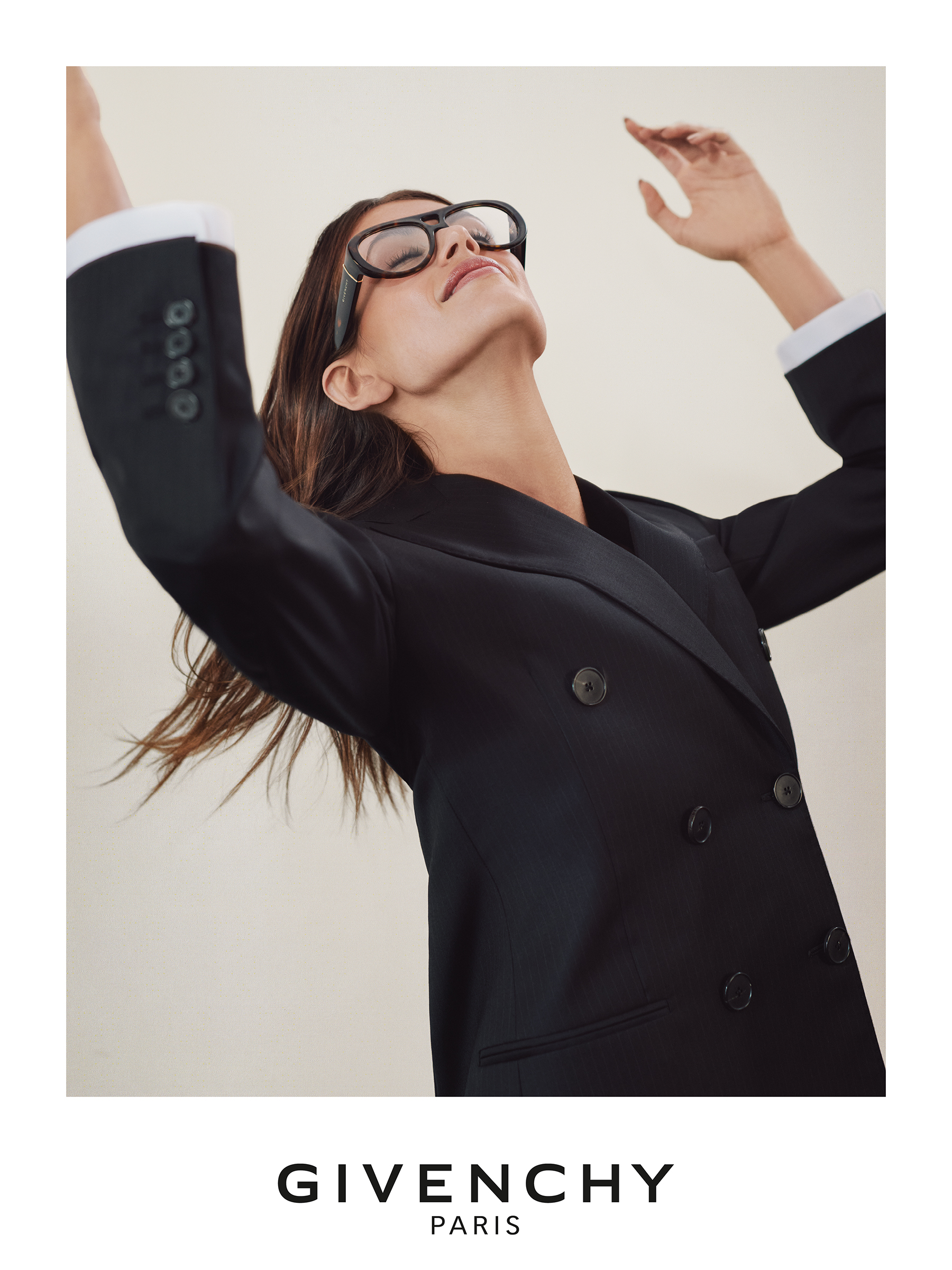 Una persona in abito nero doppiopetto e occhiali posa con le braccia alzate davanti a uno sfondo monocromatico. Logo Givenchy sul lato.