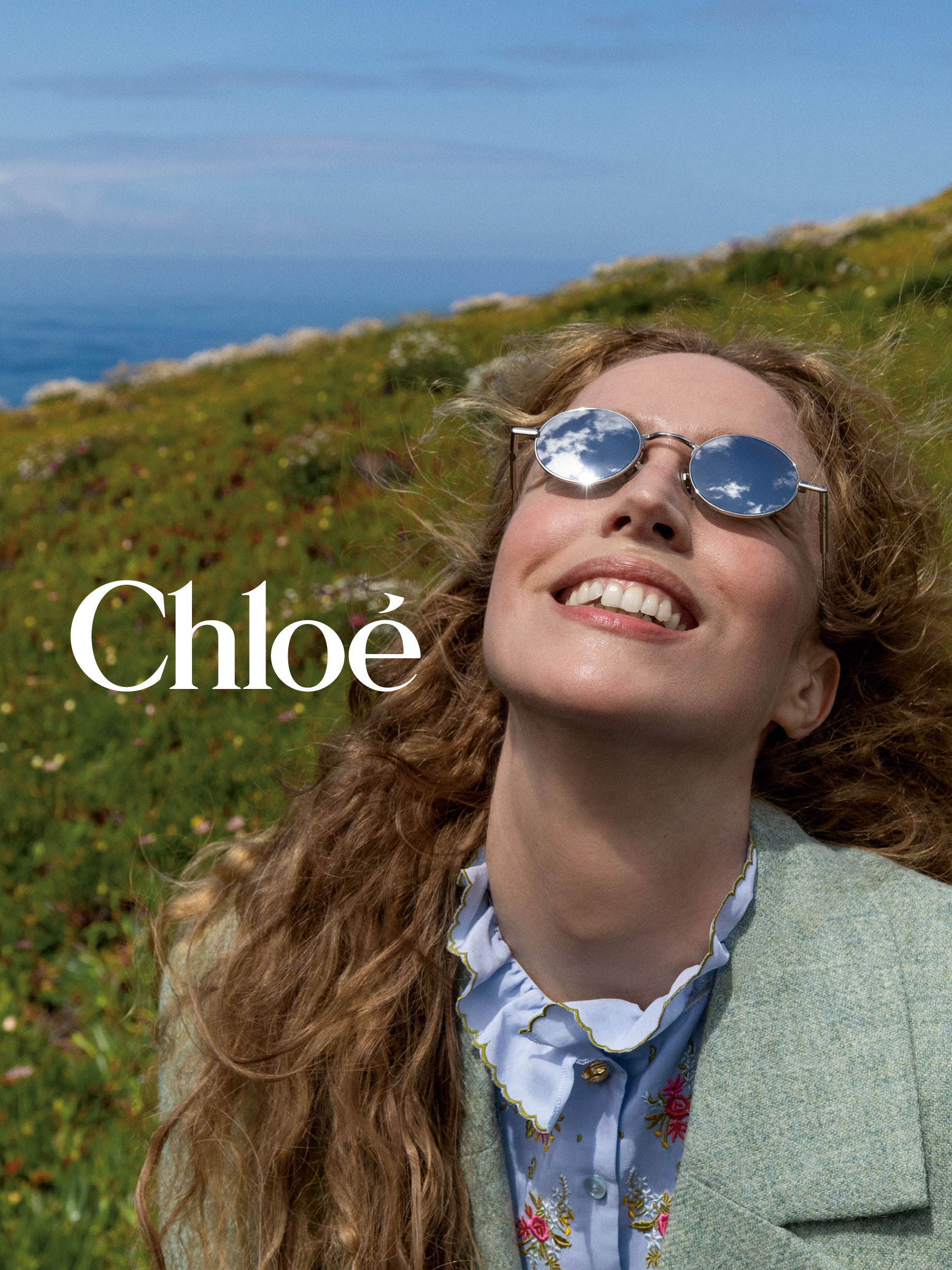 Une femme souriante, aux cheveux bouclés et portant des lunettes de soleil à verres miroirs, se tient dans un pré sous un ciel bleu. Le texte indique « Chloé ».