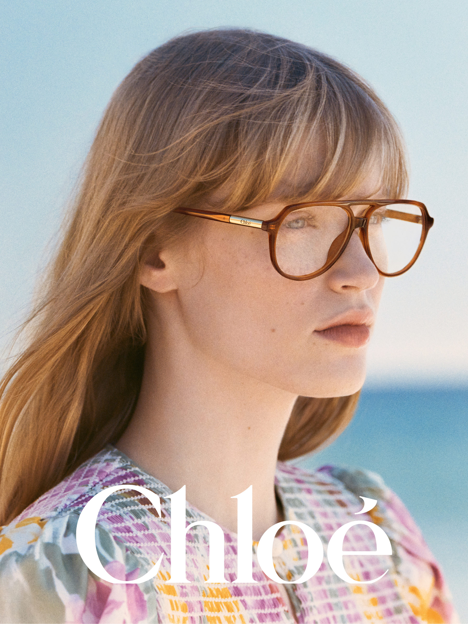 Žena s dlouhými vlasy a velkými brýlemi, má na sobě barevný vzorovaný top. Rozmazané pozadí pláže.