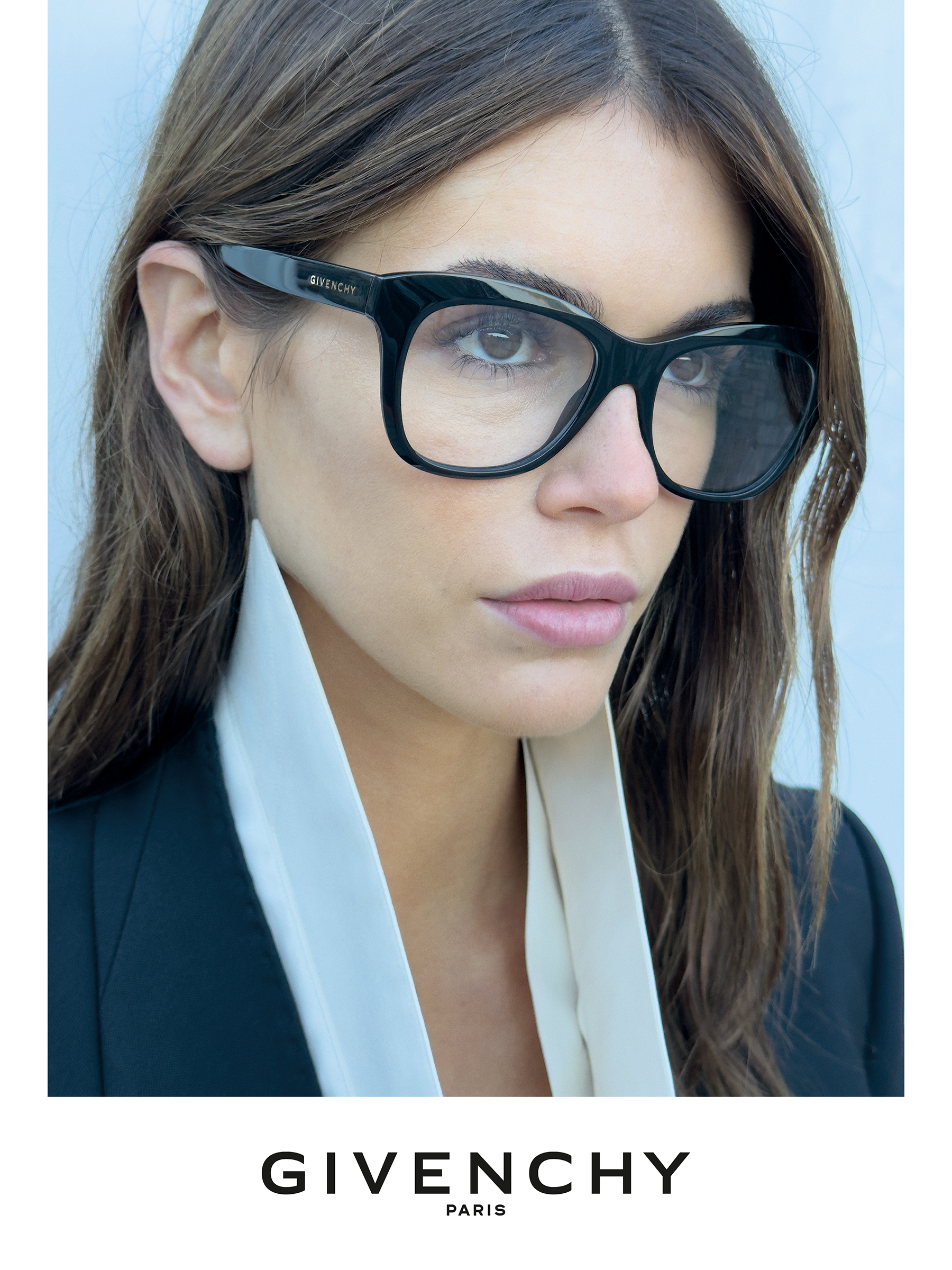 Une femme portant de grandes lunettes Givenchy noires et aux longs cheveux bruns, vêtue d'une chemise blanche à col et d'un blazer noir, devant un fond clair.