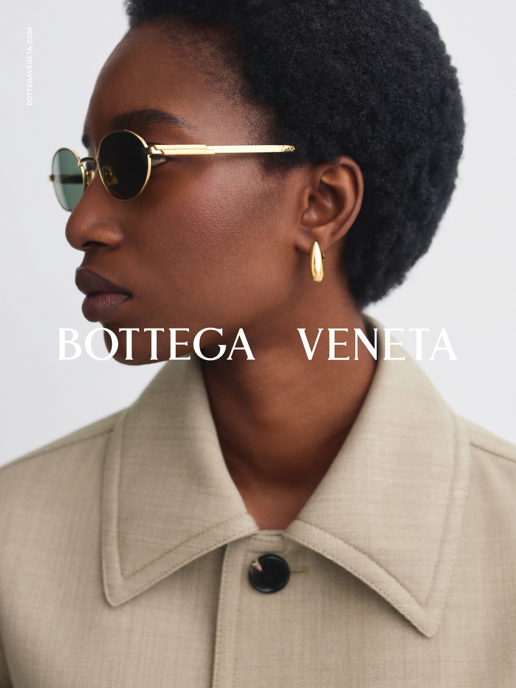 Osoba s kulatými slunečními brýlemi a zlatými kruhovými náušnicemi, oblečená v béžové bundě s velkým límcem s nápisem „Bottega Veneta”.